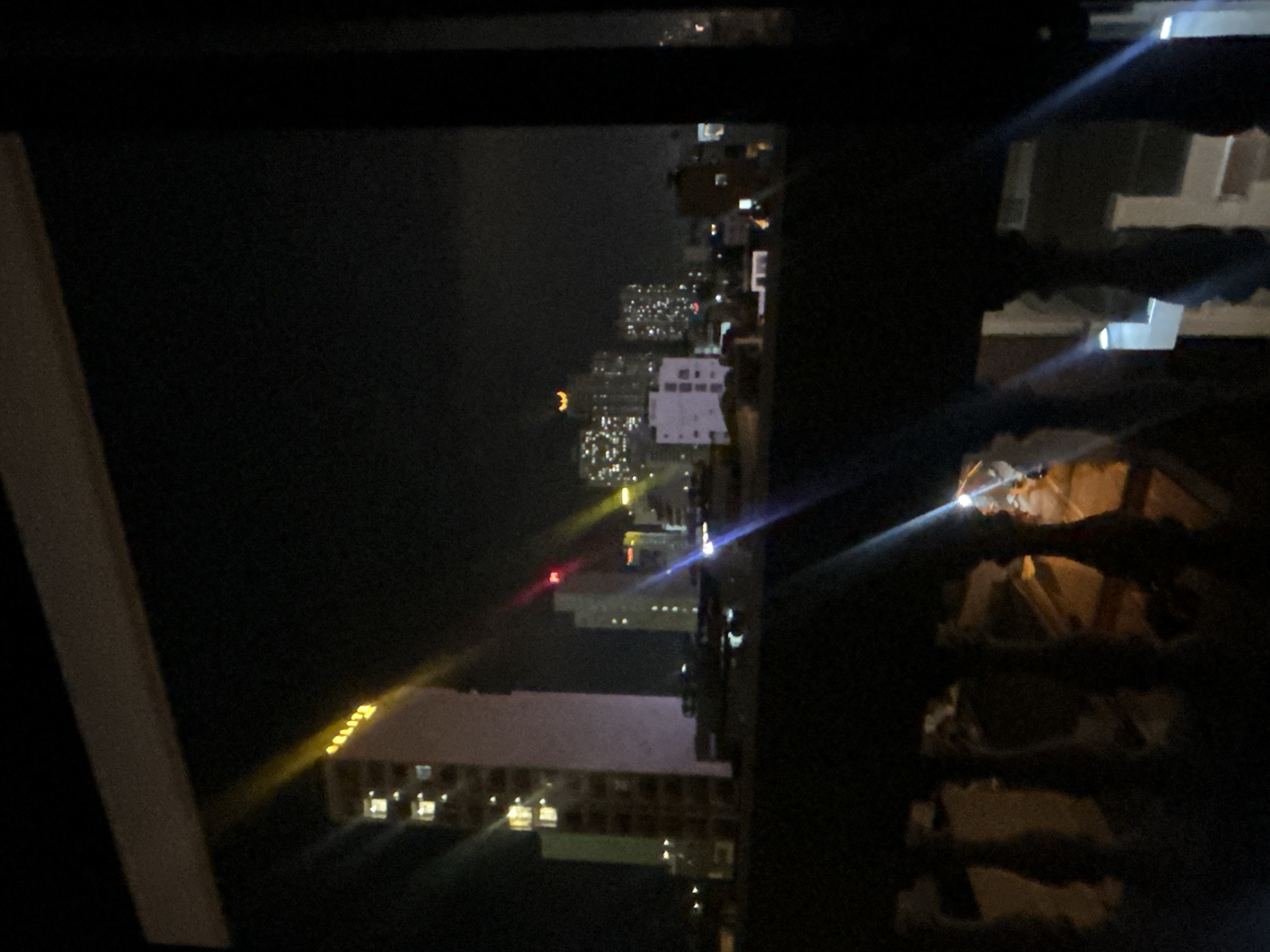 The image shows a night view of Bắc Nha Trang, Vietnam, from a high vantage point, possibly a building. The skyline is visible with various buildings illuminated by artificial lights. Some buildings are taller than others, creating an uneven cityscape. The background is dark, suggesting a clear night sky. In the foreground, a window frame or part of a building structure is visible, adding depth to the image.