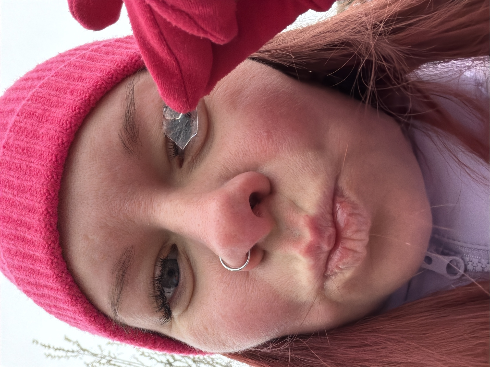 A close-up, upside-down portrait shows a person with fair skin wearing a pink knit hat and holding a small, shard-like object near their right eye. The person has red hair and is wearing a silver nose ring. A red-gloved hand is partially visible above the shard. The background is blurred, suggesting an outdoor setting with glimpses of white sky and bare branches. The zip of a white or light lavender jacket is visible on the right. The location of the image in the city of Chaikovsky, Russia cannot be determined from the image.