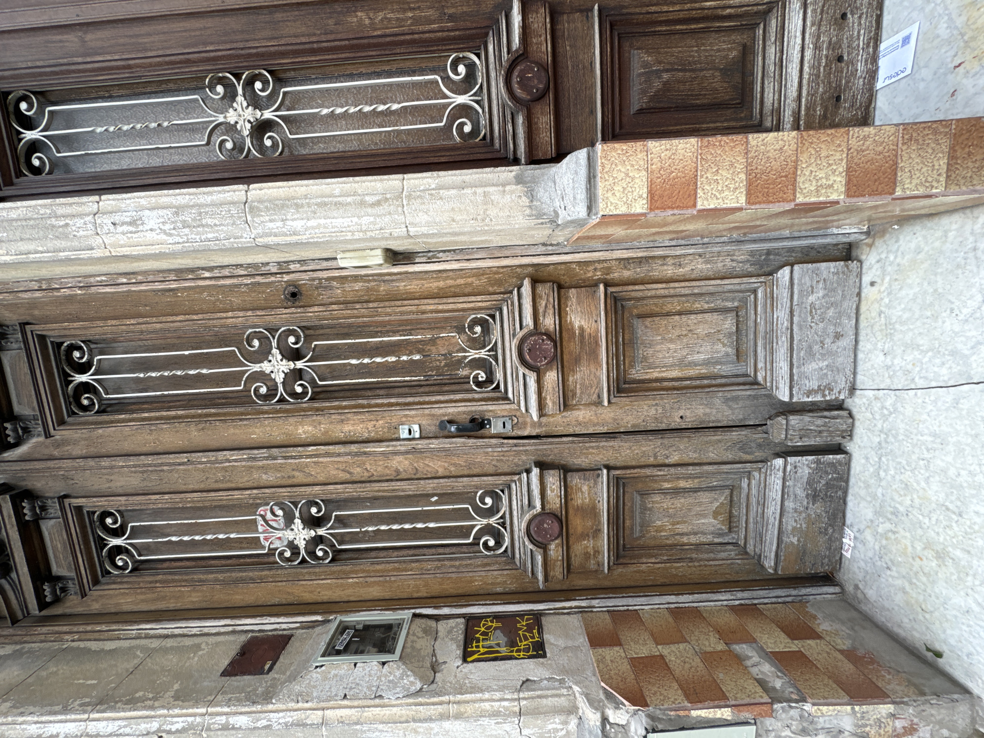 A close-up, slightly tilted shot shows a weathered, wooden double door. Each door section features a decorative metal grille over what may have been glass panels. The wood is aged with visible wear and tear. A door handle is visible on the middle section. Below the door, the wall shows signs of age and decay with visible cracks and tilework on the bottom edge. The location is Buenos Aires, Argentina.