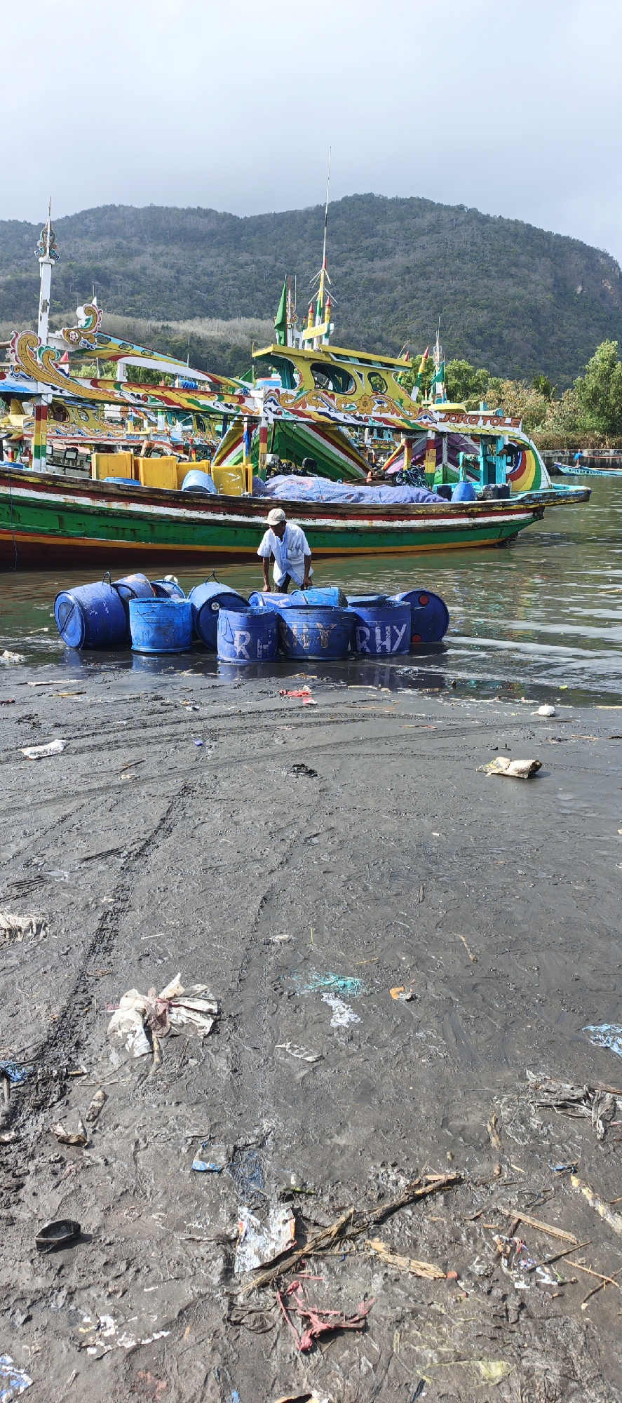 In Puger, Indonesia, a man in a white shirt and hat stands near several blue barrels, some of which have writing on them, including "RHY". The barrels are partially submerged in murky water on a muddy shore. In the background, a colorful fishing boat with elaborate decorations is moored. The sky is overcast, and a forested hill is visible behind the boat. The shore is littered with debris, including plastic and wood.