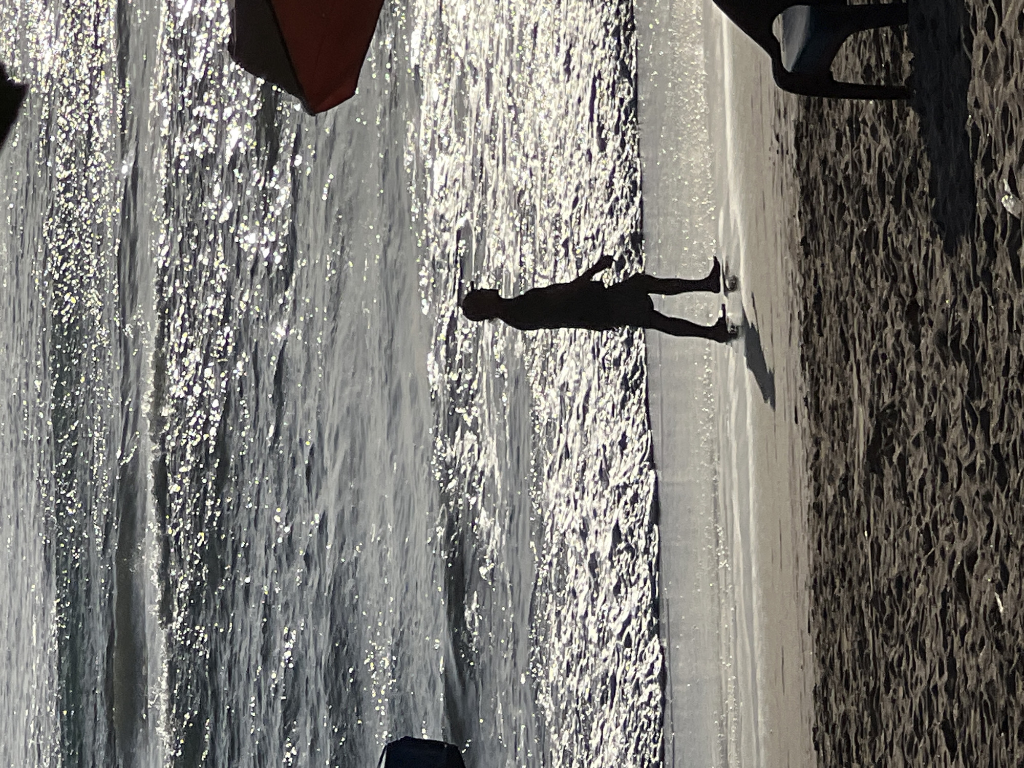 A silhouetted man walks along the shoreline of a beach. The man is wearing shorts and a cap. The sun reflects off the water, creating a shimmering effect. Part of an orange umbrella and a beach chair are visible in the foreground. The sand is dark and textured. The scene is likely the beach in Kuta, Indonesia, known for its surf and sunsets.