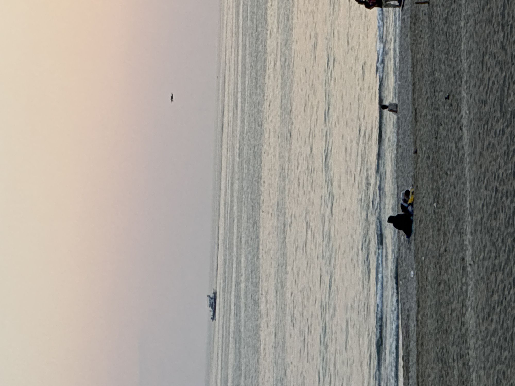 The image shows a view of the sea and a beach in Alanya, Turkey. The sea is calm with gentle waves and appears to be under a soft, hazy sky, suggesting either early morning or late afternoon light. A small boat is visible on the water in the distance.
On the beach, several people are present. One person is sitting down, dressed in black. Another person, wearing a yellow and blue outfit, is standing next to them, possibly interacting with the seated individual. The beach itself appears to be a promenade or a paved area adjacent to the sand, with a textured surface. The water meets the shore with a gentle surf line. In the far distance, a lone figure is visible against the pale sky. The overall impression is one of a peaceful seaside scene. No visible text is present in the image.