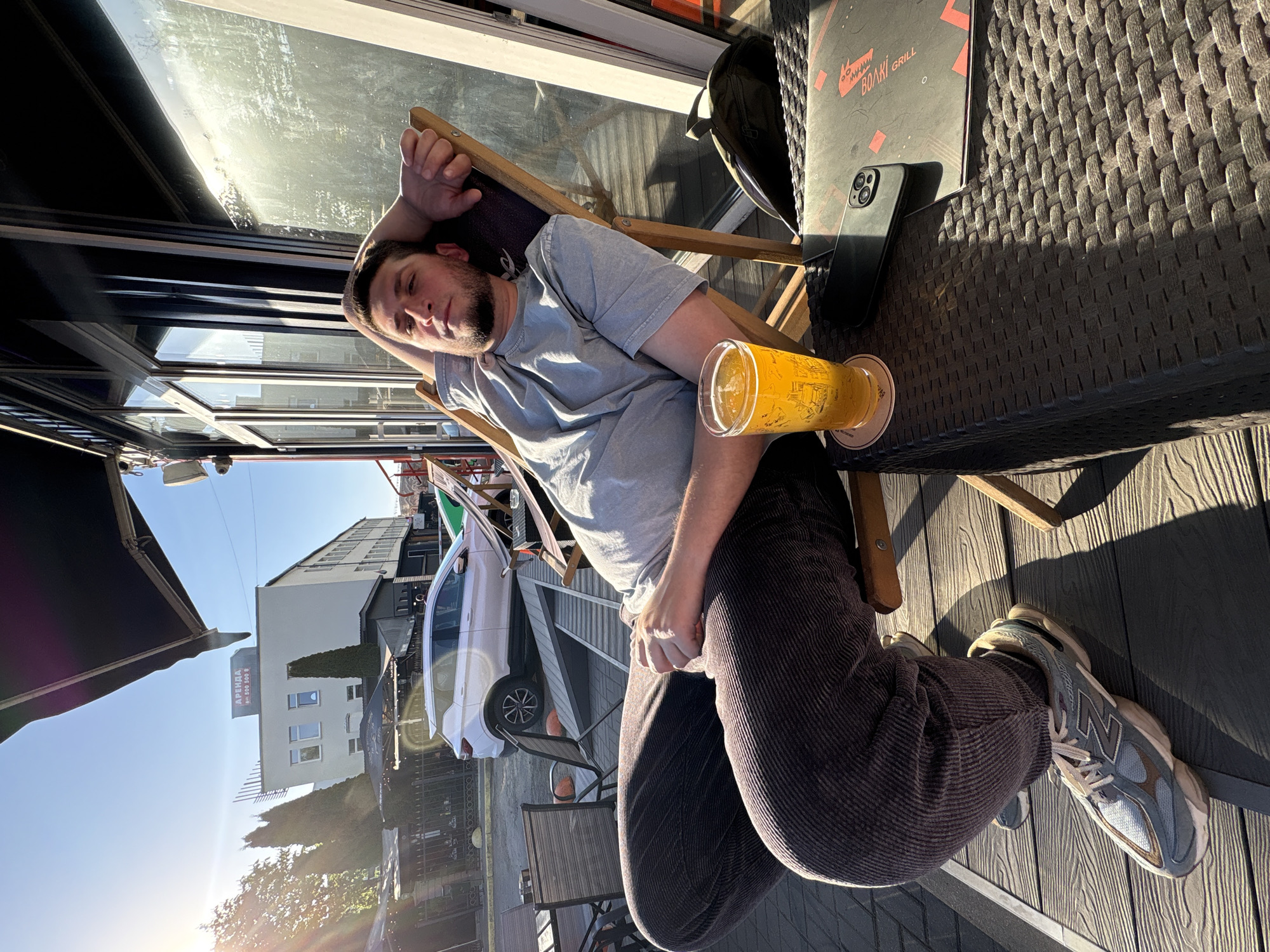 A man is reclining in a wooden chair at an outdoor cafe. He's wearing a grey t-shirt and brown corduroy pants and has a full glass of light-colored beer on the table next to him. A black menu and a cell phone are also on the table. In the background, a white building with a sign that reads "АРЕНДА" in Cyrillic script is visible, along with a white car parked outside. The cafe appears to be in Minsk, Belarus based on the Cyrillic writing. The sun is shining, suggesting it's daytime.
