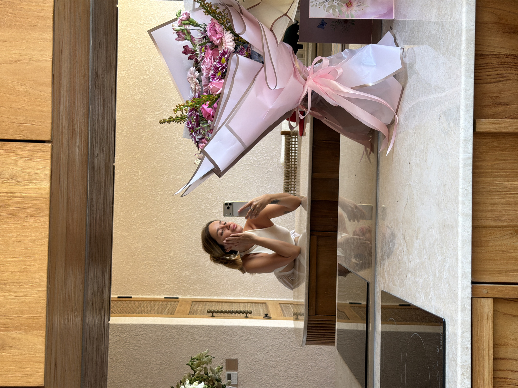 A mirrored image depicts a woman in a bathroom with a bouquet of flowers. The woman, holding a mobile phone, is wearing a light-colored top and bottoms. The bouquet, wrapped in pink and white paper with pink ribbons, is positioned near the top of the mirror. The bathroom features wood paneling and a beige-colored wall. Based on known information, this scene could be located in Mengwi, Indonesia.