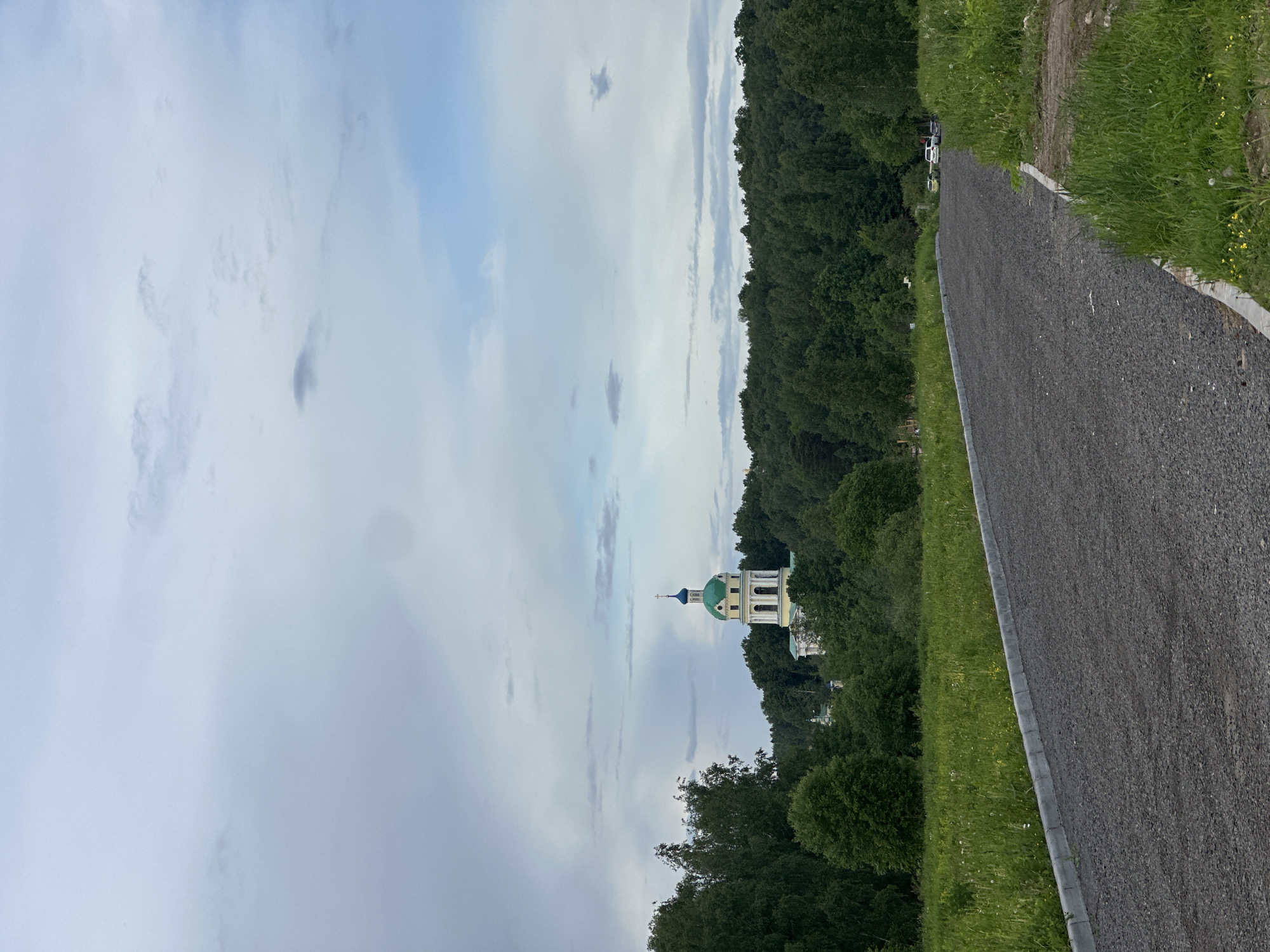 The image captures a view in Fryazino, Russia, featuring a paved pathway lined with greenery on the right. To the left of the pathway, a dense forest area extends into the background. The skyline is dominated by a cloudy sky, with a distinct church structure with a green dome and a cross on top rising above the trees, indicating a religious or historical site within the landscape.