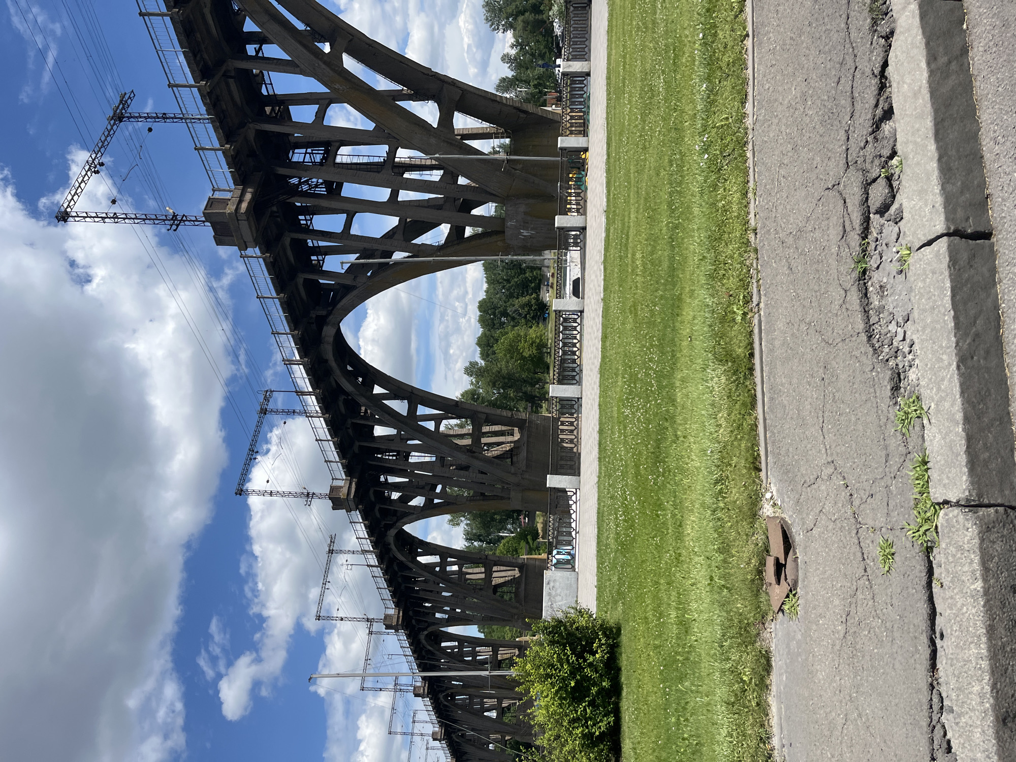 The image shows a large concrete bridge with intricate lattice-like structures against a bright blue sky with scattered white clouds. Power lines and their supporting structures are visible beneath and alongside the bridge. To the right of the bridge, a manicured green lawn is bordered by a paved walkway with a decorative railing. Beyond the walkway, trees form a dense line of foliage. The foreground features a section of cracked asphalt road with a damaged manhole cover. The scene appears to be set during the daytime under clear weather conditions. The overall impression is one of industrial infrastructure integrated with a well-maintained public space. The location is identified as Dnipro, Ukraine.