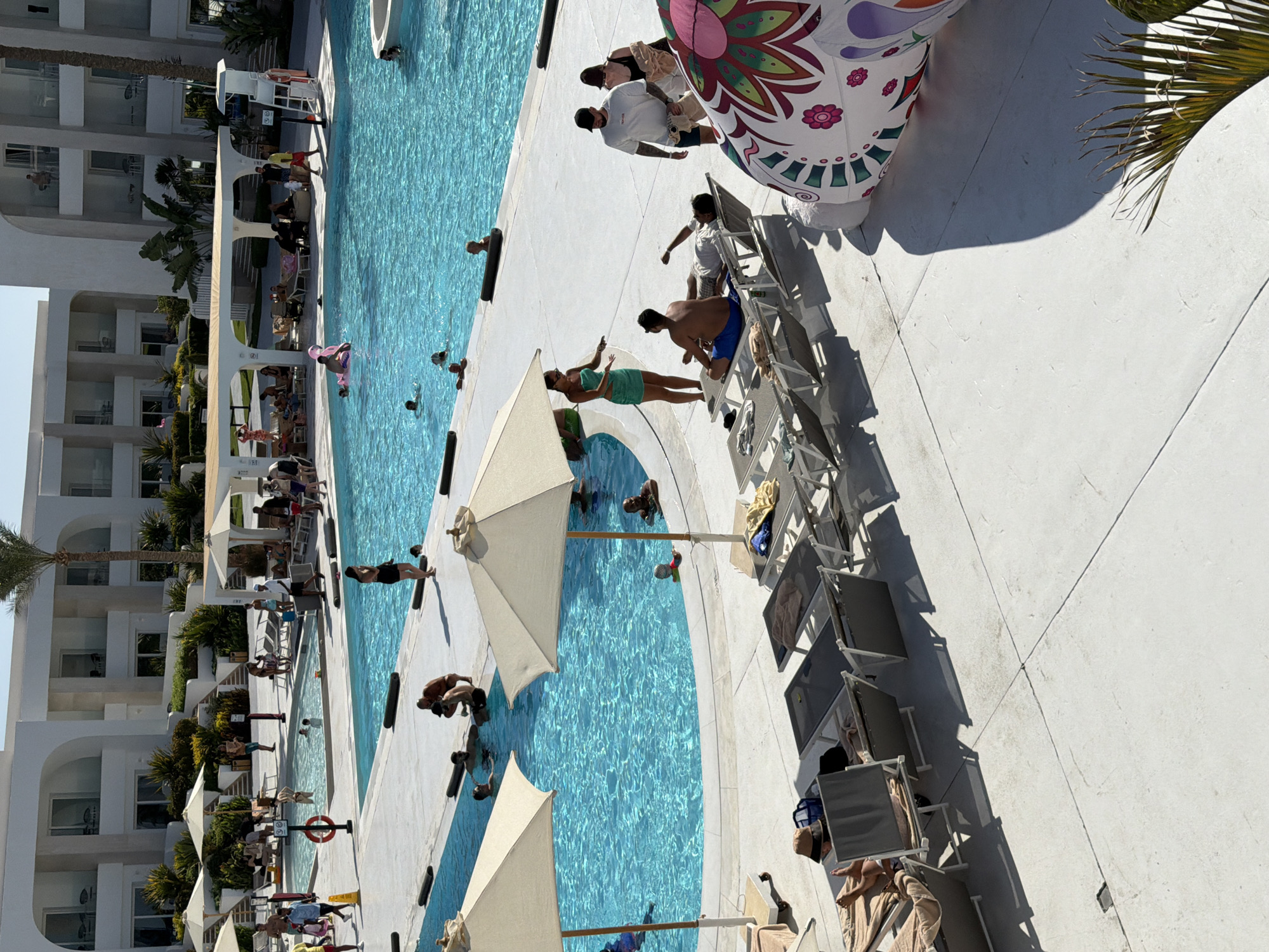 The image depicts a resort in Sharm El Sheikh, Egypt, featuring a large, blue swimming pool surrounded by lounge chairs and umbrellas. Several people are visible in and around the pool. In the foreground, lounge chairs are set up on a white concrete patio. Some chairs are occupied by individuals relaxing, while others are empty. Two large, cream-colored umbrellas provide shade over some of the chairs.
In the pool, various individuals are swimming and interacting. Some are floating, while others are actively swimming. A person is seen on a pink inflatable float. The water is a vibrant blue, reflecting the clear sky.
The background shows a multi-story building, characteristic of resort architecture, with balconies and windows. Lush green vegetation and palm trees are interspersed around the buildings and pool area. There is also a lifeguard stand with a ladder and a red life preserver. A large, inflatable decoration with colorful floral patterns is partially visible in the lower right corner of the image. The overall scene conveys a sense of leisure and recreation at a tropical resort.