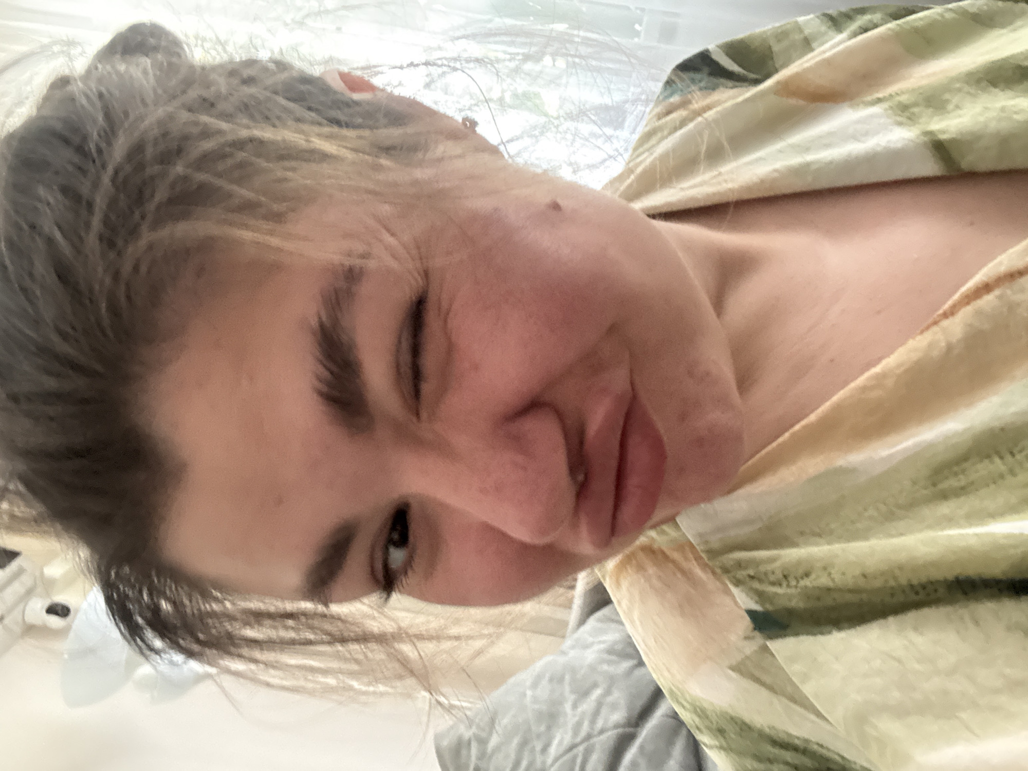 A close-up selfie shot shows a person with fair skin and dark brown hair pulled back in a loose bun. The person is winking one eye and has pursed lips. The person is wearing a light-colored shirt with a green and beige pattern. The background is blurry, but shows a window and possible room interior. There is no verifiable information to determine the location of this image as Votkinsk, Russia.