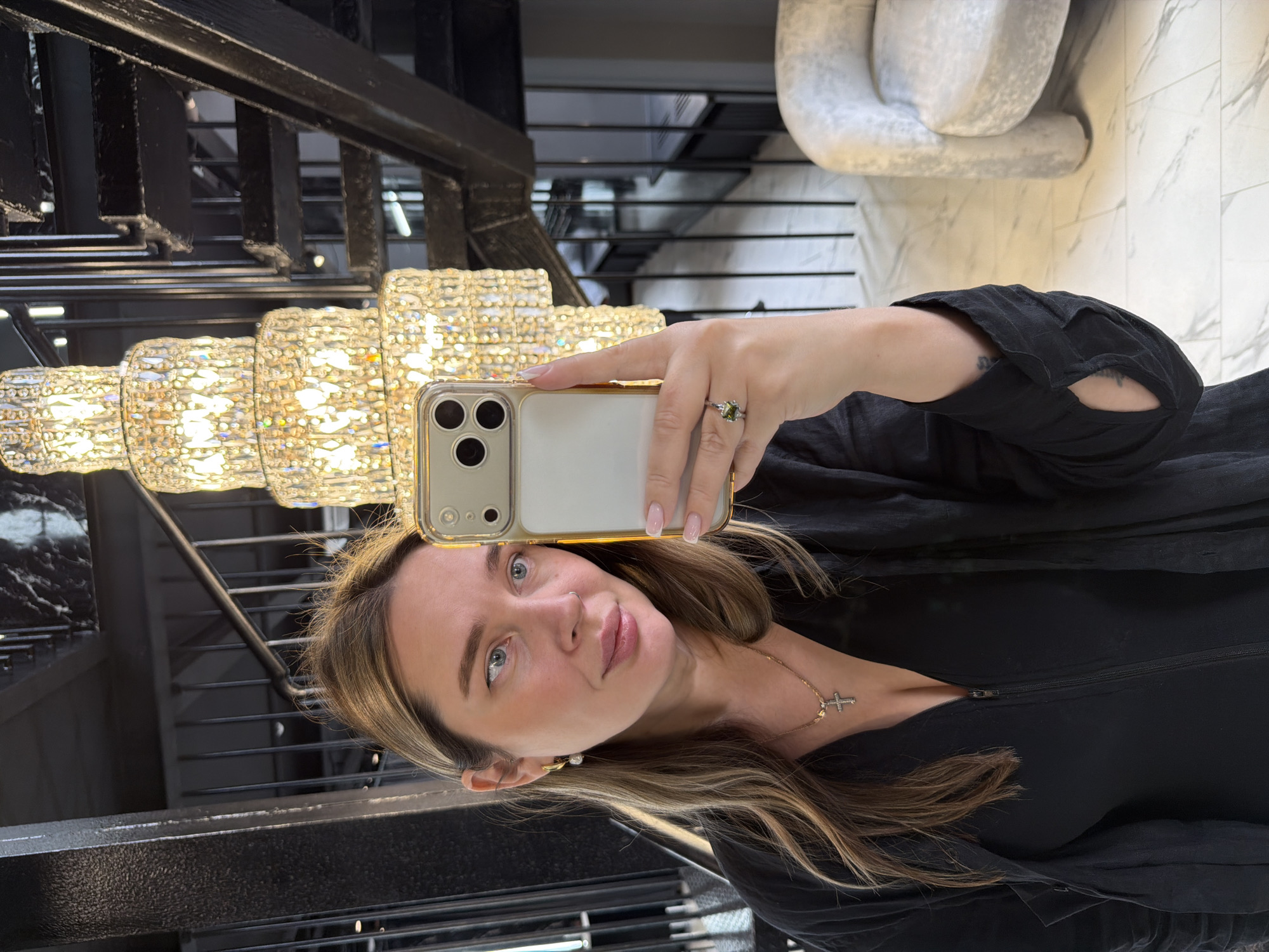 An indoor mirror selfie captures a woman posing in a modern, luxurious setting, likely in Moscow, Russia. The woman, with medium-length brown hair and subtle highlights, blue eyes, and light makeup, looks directly into the camera with a neutral to slight smile. She wears a nose piercing, a gold necklace with a cross pendant, and small, pearl-like earrings. Her left hand, adorned with light pink nail polish and a ring featuring a green stone, holds a gold-cased smartphone with a triple camera setup. She is dressed in a black top or dress, covered by a black, loose-fitting outer garment, revealing tattoos on her left arm.
The reflected background reveals a sophisticated interior. Dominating the left side is a multi-tiered, opulent chandelier-like light fixture, composed of numerous glittering clear or crystalline elements, emitting a warm glow. Behind it, a dark, possibly metallic, staircase or balcony railing with horizontal bars ascends. To the right, a large, abstract sculpture or piece of furniture in a light, textured material rests on what appears to be a white marble-patterned floor or wall. The scene suggests an upscale lobby, commercial space, or a grand residence. No visible text is present in the image.