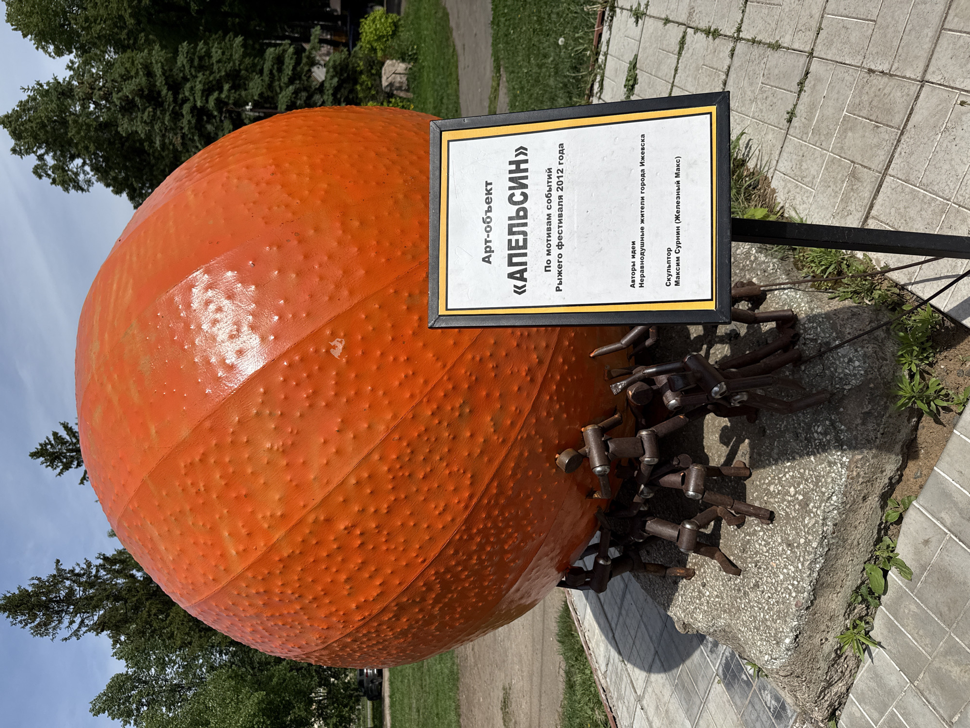 The image shows an art installation, titled "Orange" ("Апельсин" in Russian), located in the city of Izhevsk, Russia. The installation features a large, orange-colored sphere resembling an orange, placed on a stone base. The sphere is textured with small bumps, suggesting the peel of an orange. Metal rods protrude from underneath the sphere, appearing like roots or supporting structures. A sign next to the orange identifies the artwork and provides information about its creation. It states that the artwork is based on the events of the "Ginger Festival" of 2012. The sign also credits the idea to caring residents of Izhevsk and names Maxim Surnin (Железный Макс) as the sculptor. The installation is set in an outdoor environment, with trees and a paved walkway visible in the background.