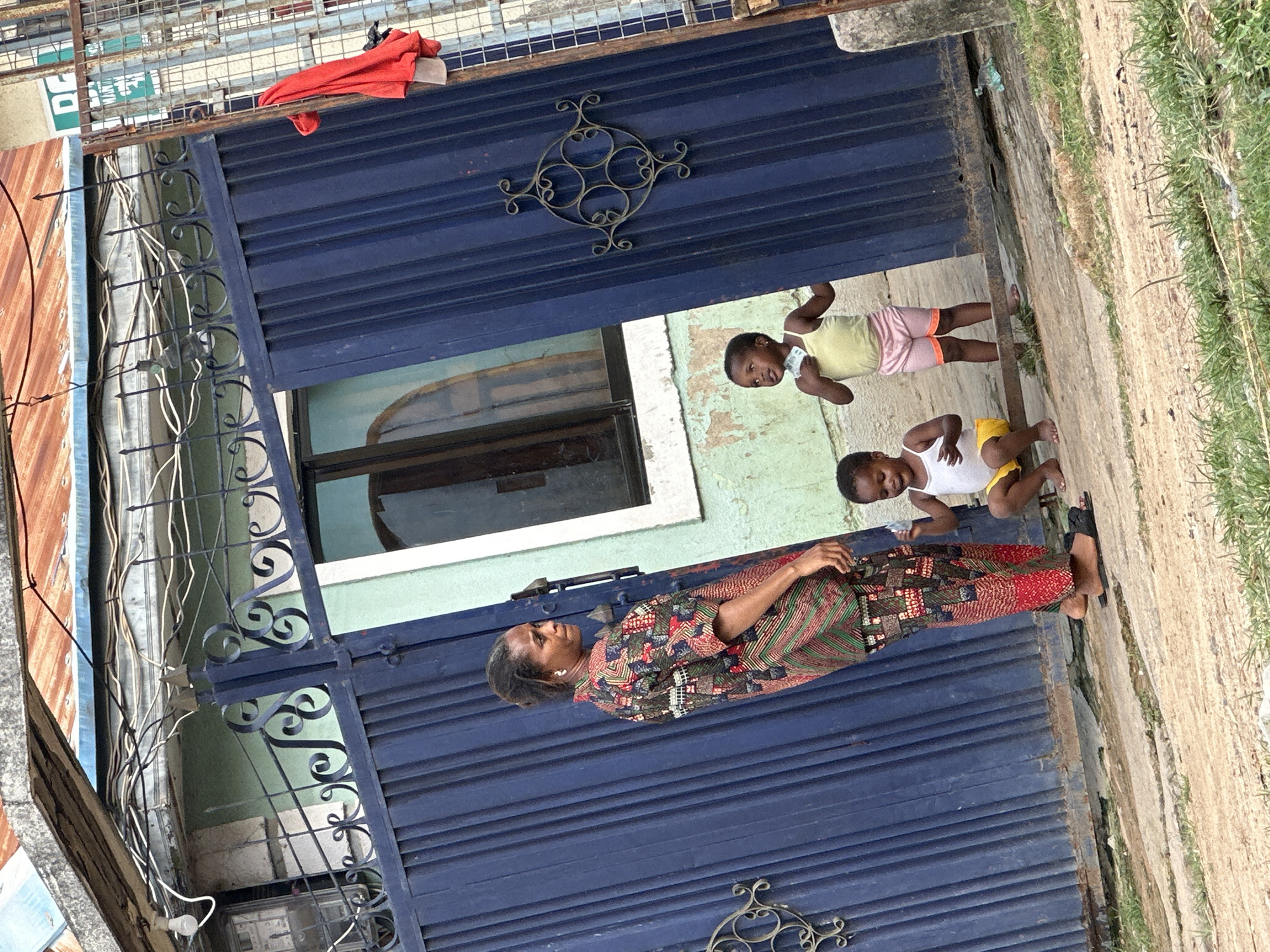 In Narayi, Nigeria, a woman in a vibrantly patterned traditional outfit stands to the left of a tall, dark blue metal gate. Her hair is pulled back, and she wears white earrings. In front of the gate, three young children are visible. The child on the far left, wearing a white tank top and yellow shorts, is crouched down. To their right, a child in a yellow tank top and pink shorts stands, holding what appears to be a small stack of bills. Further to the right, a child in a white tank top and yellow shorts is also crouched, looking upwards. The gate features decorative metalwork and is slightly ajar, revealing a glimpse of a window with a curved top. The building behind the gate has light green walls with peeling paint and a rough, textured surface. Above the gate, exposed electrical wires run across the frame, supported by a rusty metal structure and a weathered concrete overhang. A piece of red fabric is draped over a metal fence to the right of the gate. The ground in front of the gate is a dirt and concrete path with sparse green grass growing in patches.