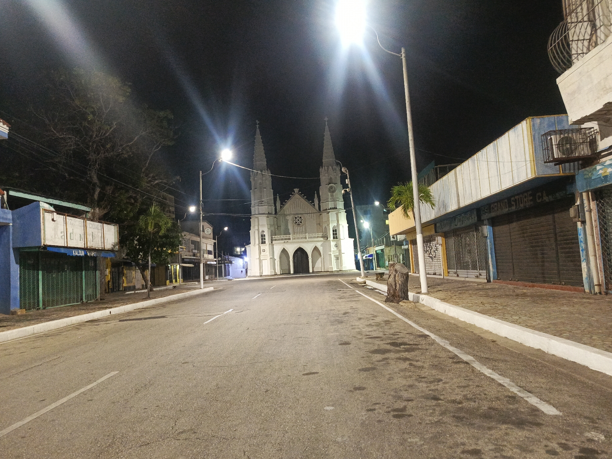 This image depicts an empty street at night in Juan Griego, Venezuela, with a prominent white church as the central focal point. The church features twin spires and arched doorways, illuminated by streetlights. The street itself is paved and has white lane markings. On either side of the street are closed storefronts with metal roll-down shutters, suggesting a commercial area. Some of the storefronts have faded signage, including what appears to be "GRAND STORE CA". A tree stump stands on the right side of the road, and a palm tree is visible to the left of the church. The overall scene is quiet and deserted, bathed in artificial light against the dark night sky.