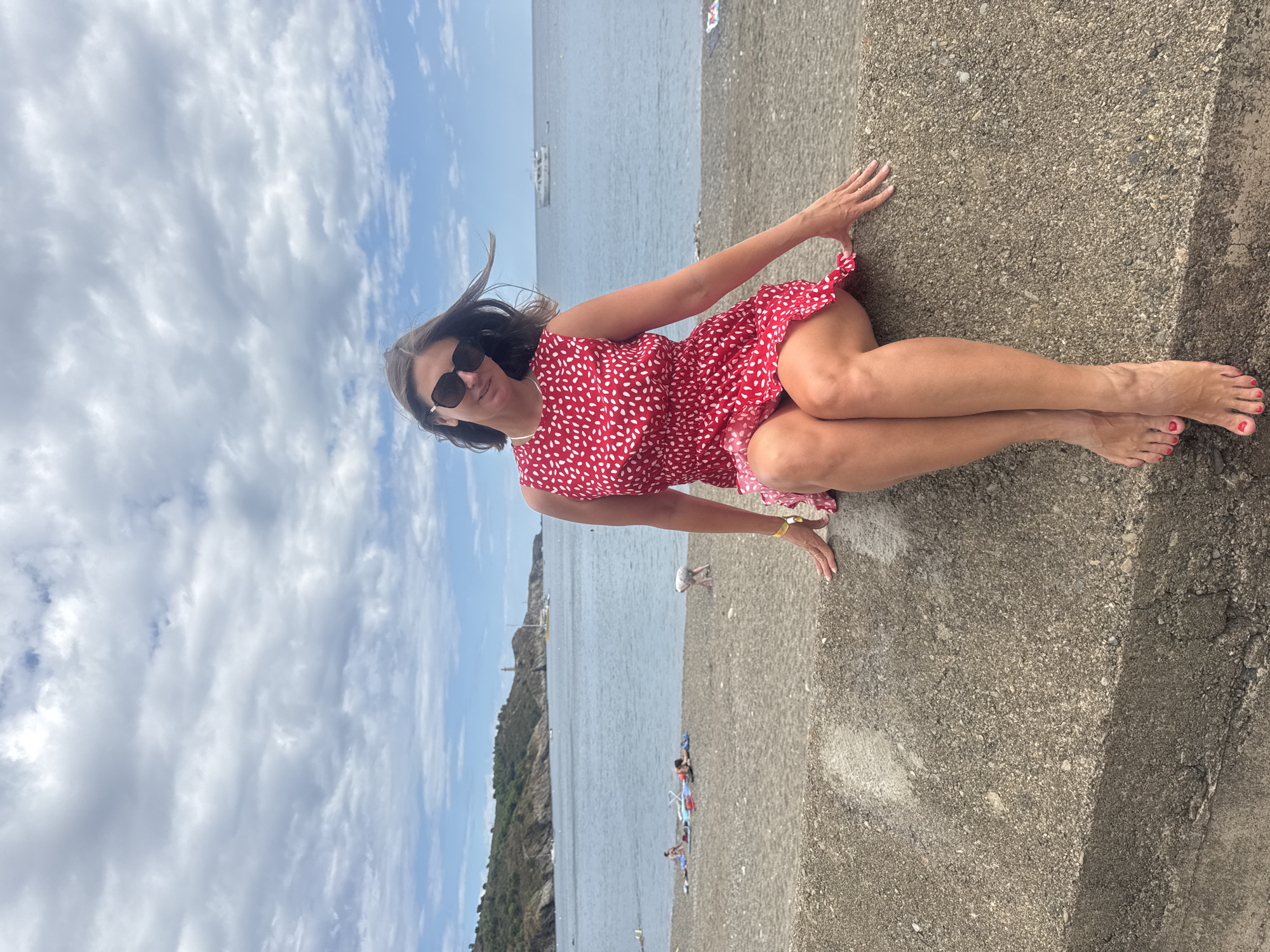 A woman in a red and white polka dot dress and sunglasses is sitting on a concrete barrier at a beach in Port-Vendres, France. She is facing the camera with her legs crossed and her hands resting on the barrier. The sea is in the background, with a few boats visible. A hilly coastline stretches along the horizon. Several people are sunbathing on the pebble beach. The sky is cloudy.