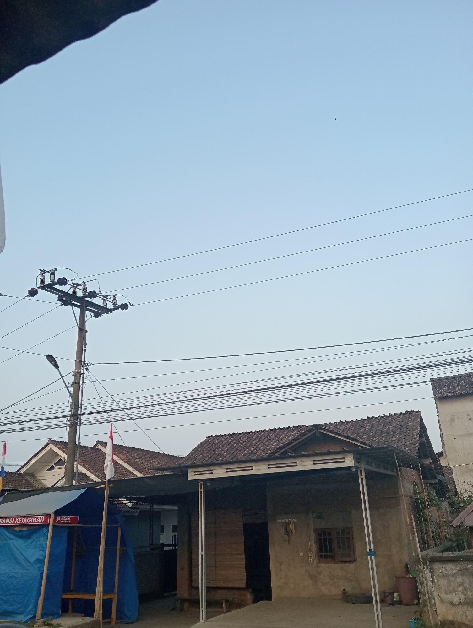 The image depicts a street scene in Pringsewu, Indonesia, under a clear sky. A utility pole with numerous wires is prominently featured on the left side of the frame, along with a streetlamp attached to it. In the foreground, a structure with a blue tarp and bamboo supports is visible, bearing the text "TRANSIT KETAGIHAN" and a logo. Adjacent to this, a house with a tiled roof and a partially open shuttered window is seen. Another house with a similar tiled roof is located behind it. A red and white flag, possibly the Indonesian flag, is planted in front of the blue-tarped structure. Power lines crisscross the sky above the buildings. The overall environment suggests a residential or commercial area in a tropical climate.