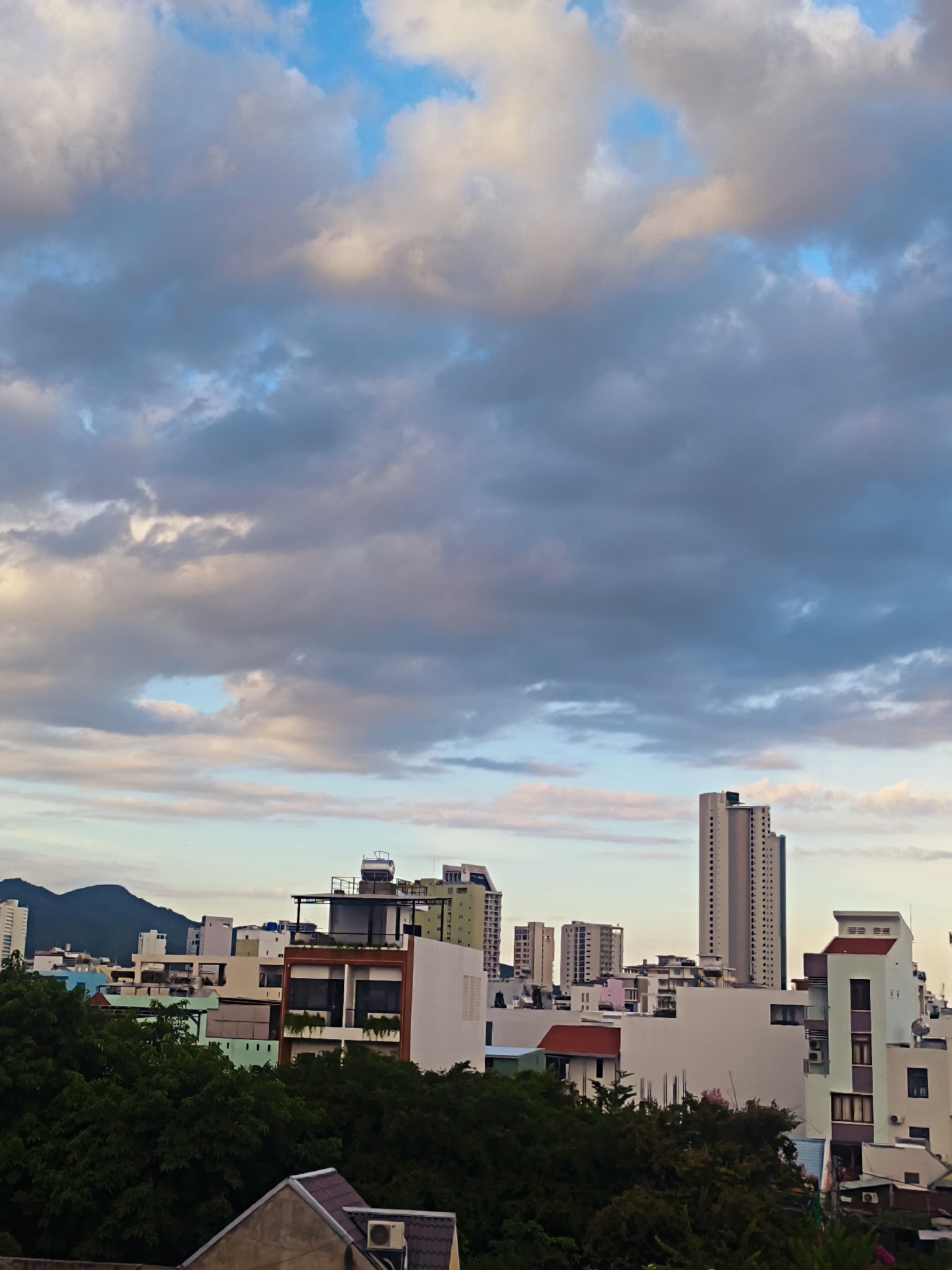 A cityscape is captured from an elevated vantage point, showcasing a dense collection of buildings against a dramatic sky. The sky is filled with a mix of fluffy white and dark gray clouds, interspersed with patches of pale blue. In the middle ground, a variety of buildings are visible, ranging from low-rise structures with red roofs to taller, modern buildings. Some buildings are white, while others display colorful facades in green and blue. Lush green trees dominate the foreground, partially obscuring the lower portions of the buildings. In the far distance, dark, undulating hills are silhouetted against the horizon. A tall, slender skyscraper with numerous windows stands out among the buildings in the middle ground, drawing the eye upwards. The overall scene suggests a view of a coastal city, possibly during the late afternoon or early evening, given the lighting and cloud formations.