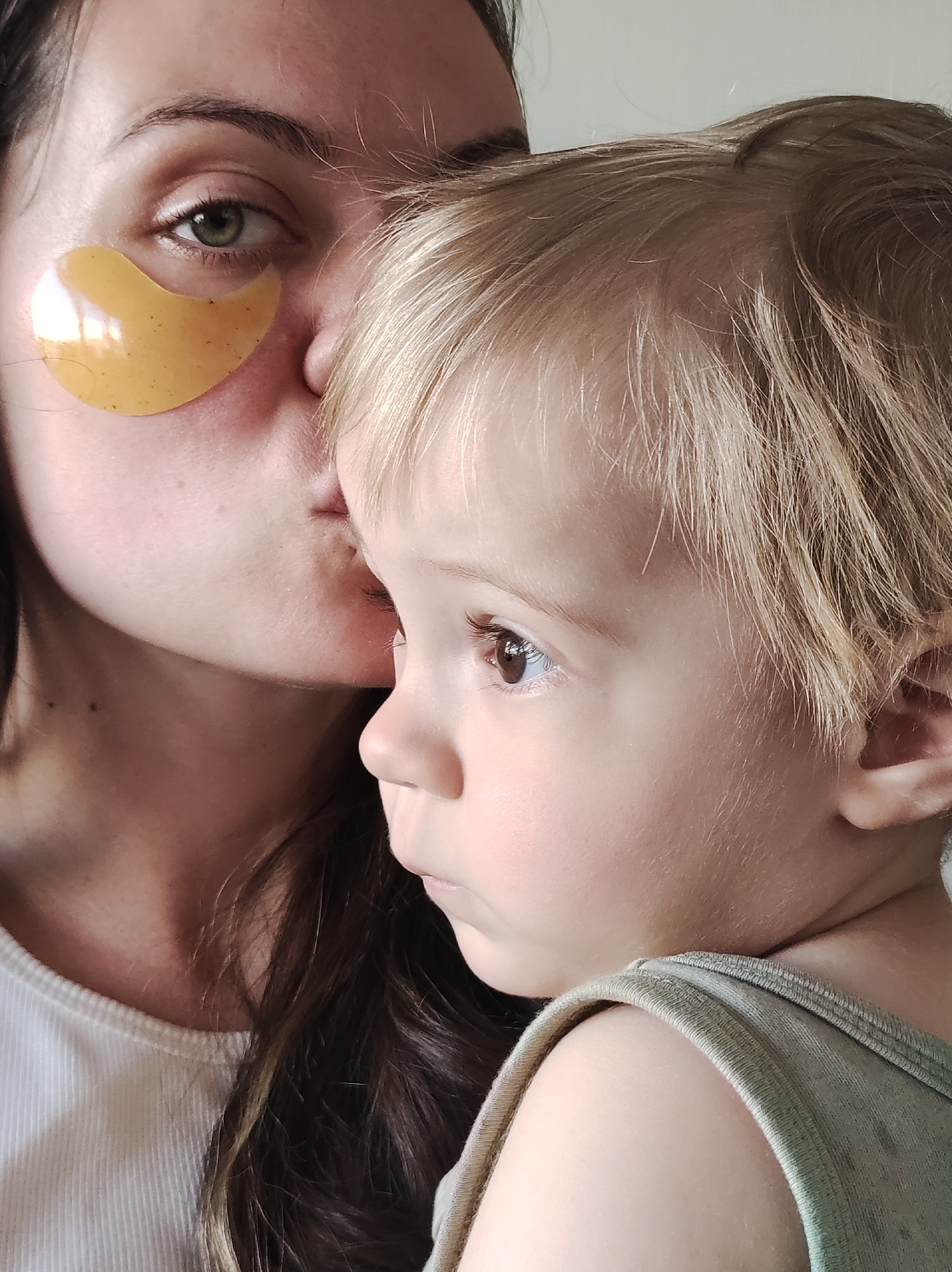A woman with dark hair and a gold under-eye patch kisses the cheek of a blonde toddler. The woman's head is tilted downwards, and her eyes are closed as she leans into the child. The toddler's head is turned away from the camera, looking to the right. The child has fine, light blonde hair and is wearing a light green sleeveless top. The background is a plain, light-colored wall.