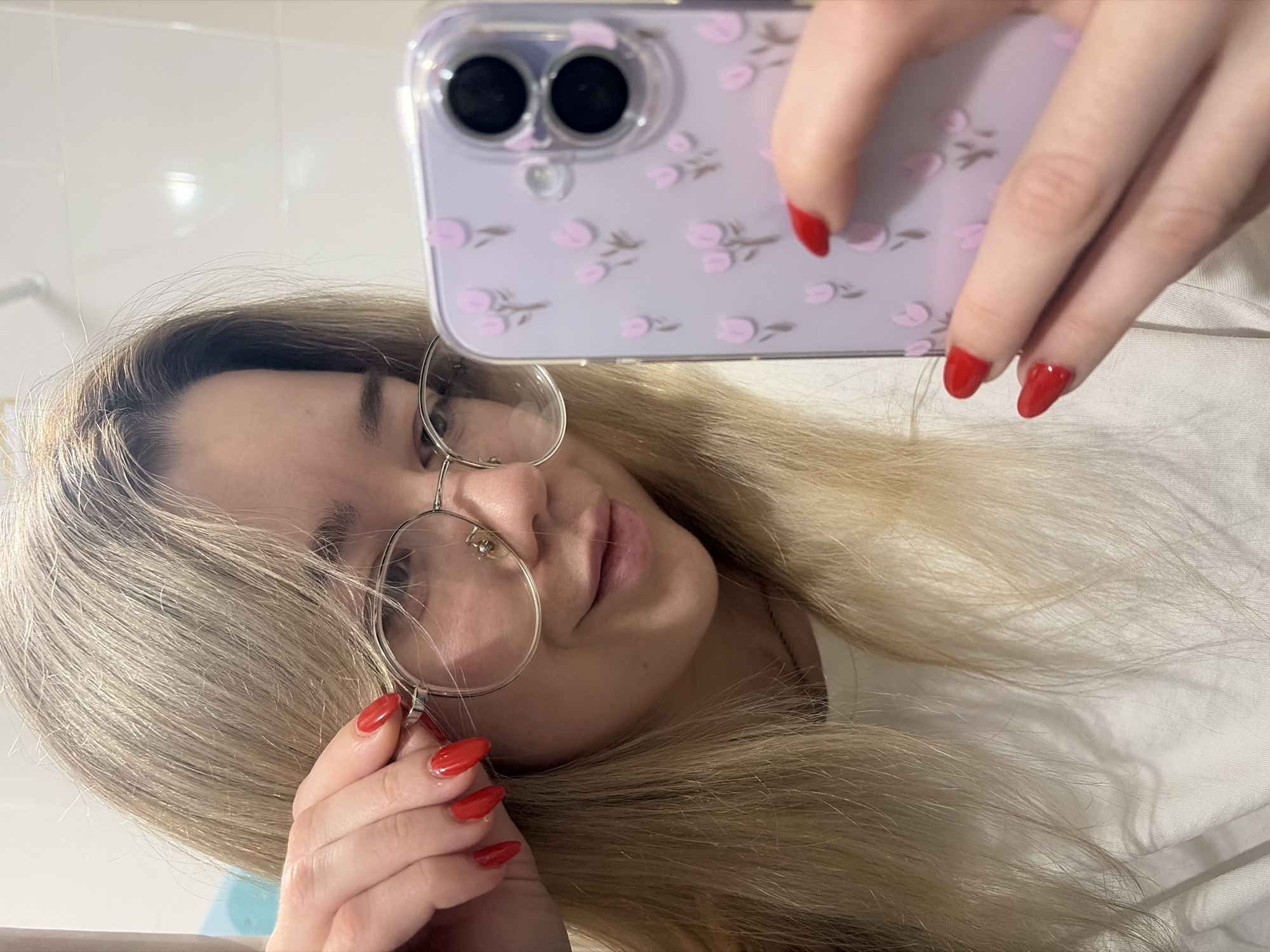 A young woman is taking a selfie in what appears to be a bathroom. She holds a purple phone case with a floral design in her right hand, obscuring part of her face. She is wearing round-rimmed glasses and has long, blonde hair. Her fingernails are painted a vibrant red. The background shows a white tiled wall. The location is not discernible from the image.