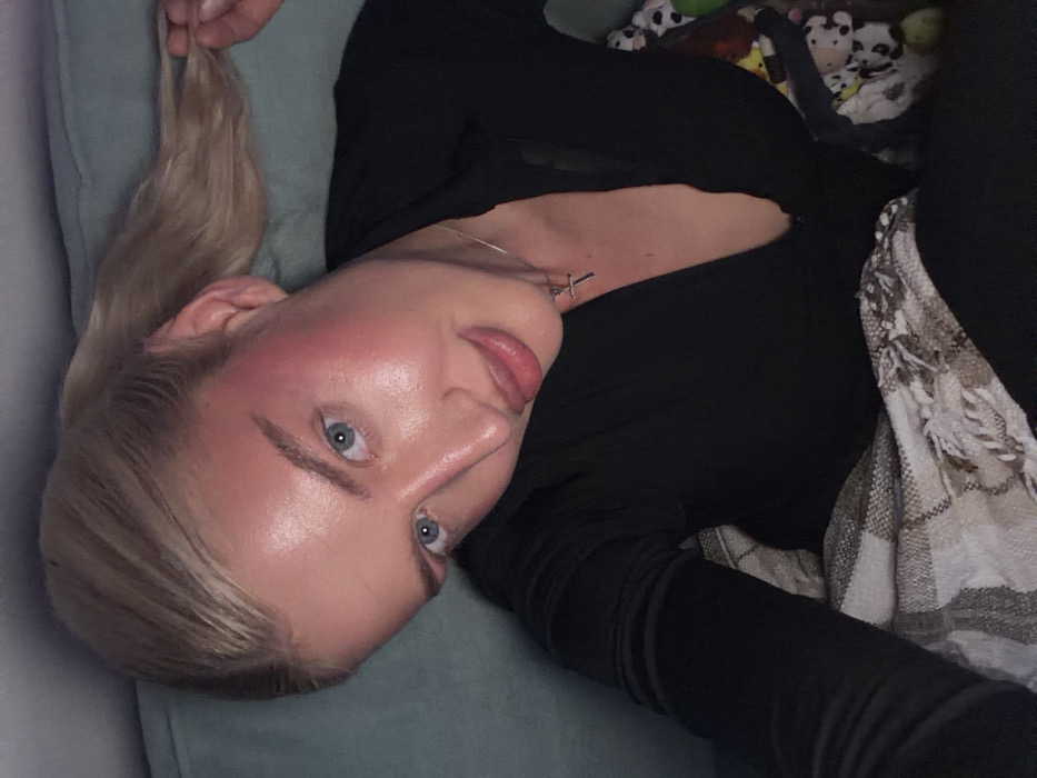 A blonde female is depicted in a close-up, slightly angled shot within an indoor setting. She has light blue eyes and fair skin exhibiting prominent reddish blush across her cheeks and forehead. Her blonde hair is pulled back in a ponytail, with her right hand grasping a portion of it. She is dressed in a dark, long-sleeved garment featuring a V-neckline. A silver-colored chain with a small cross pendant is visible around her neck. Her mouth is closed with a slight pout. A patterned blanket with white fringe covers her lower body and left arm. Behind her, a teal upholstered surface is visible, likely part of a sofa or bed. A colorful, indistinct object is partially visible to her right. The specific location within the city of Yurlovo, Russia, cannot be determined from the visual information provided.