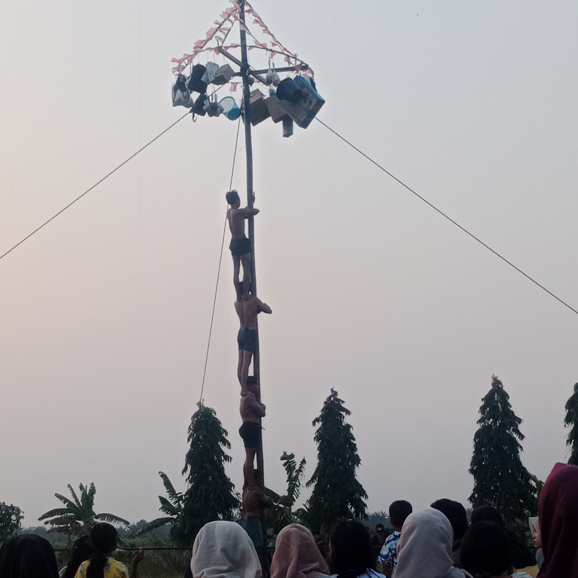 In Tegowanu Wetan, Indonesia, a group of young men are participating in a pole climbing competition. The main subject is a tall wooden pole adorned with red and white triangular flags and various objects like bags and baskets, suspended from the top. Several shirtless young men are climbing the pole, with one man standing on the shoulders of another, forming a human pyramid. Spectators, including women and children, are gathered at the base of the pole, watching the event. The environment appears to be an outdoor setting with trees and a hazy sky, suggesting it is either early morning or late afternoon.
