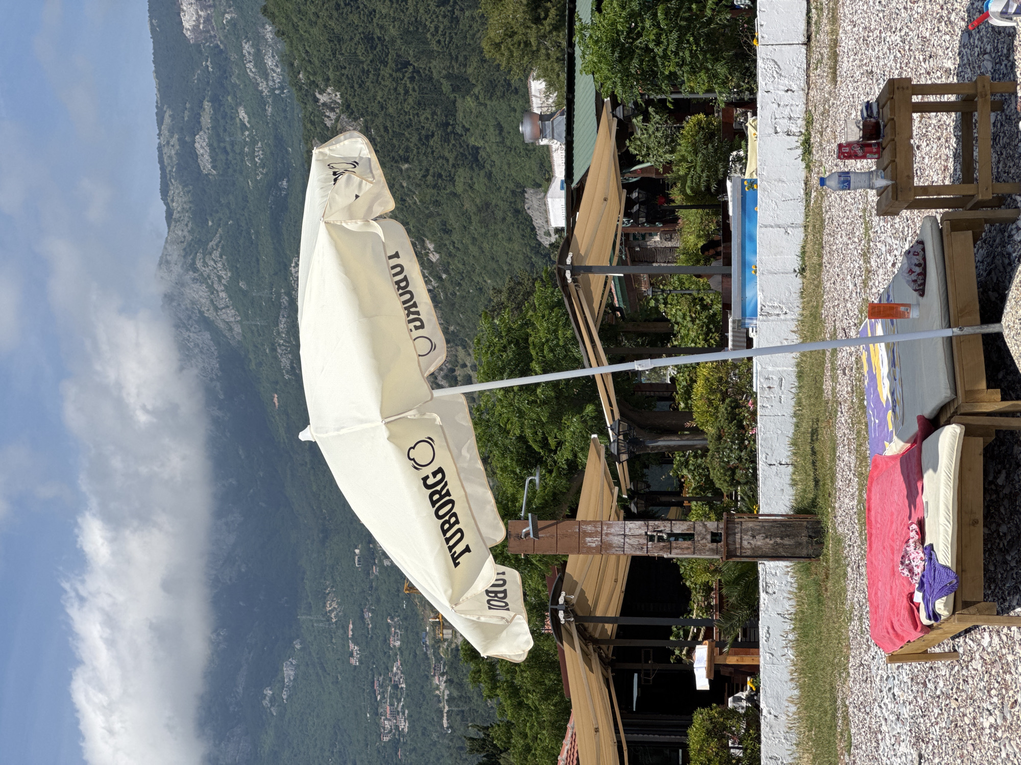 The image presents a scene in what appears to be a resort setting. There is a closed white Tuborg-branded umbrella to the left, a mountain range in the background, and resort structures on the right. A wall and some lounge chairs with towels on them are also present. The location is likely Budva, Montenegro, given the context of beach resort properties.