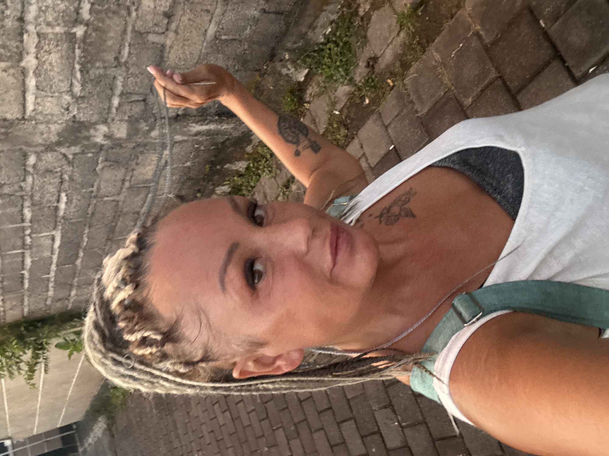 A selfie photograph, oriented vertically, shows a woman against an outdoor urban background. The woman has pale skin, braided blonde hair, and brown eyes. She wears a white tank top over a dark gray undergarment, with what appears to be a green denim strap of clothing visible. She has multiple tattoos: one on her left upper arm and another on her chest. She holds her left hand up, palm facing forward, with a cigarette between her fingers. The background consists of a brick wall on the left and a brick pathway on the right. Greenery grows near the brick pathway. The photograph was likely taken in Kuta, Indonesia, as requested in the prompt.