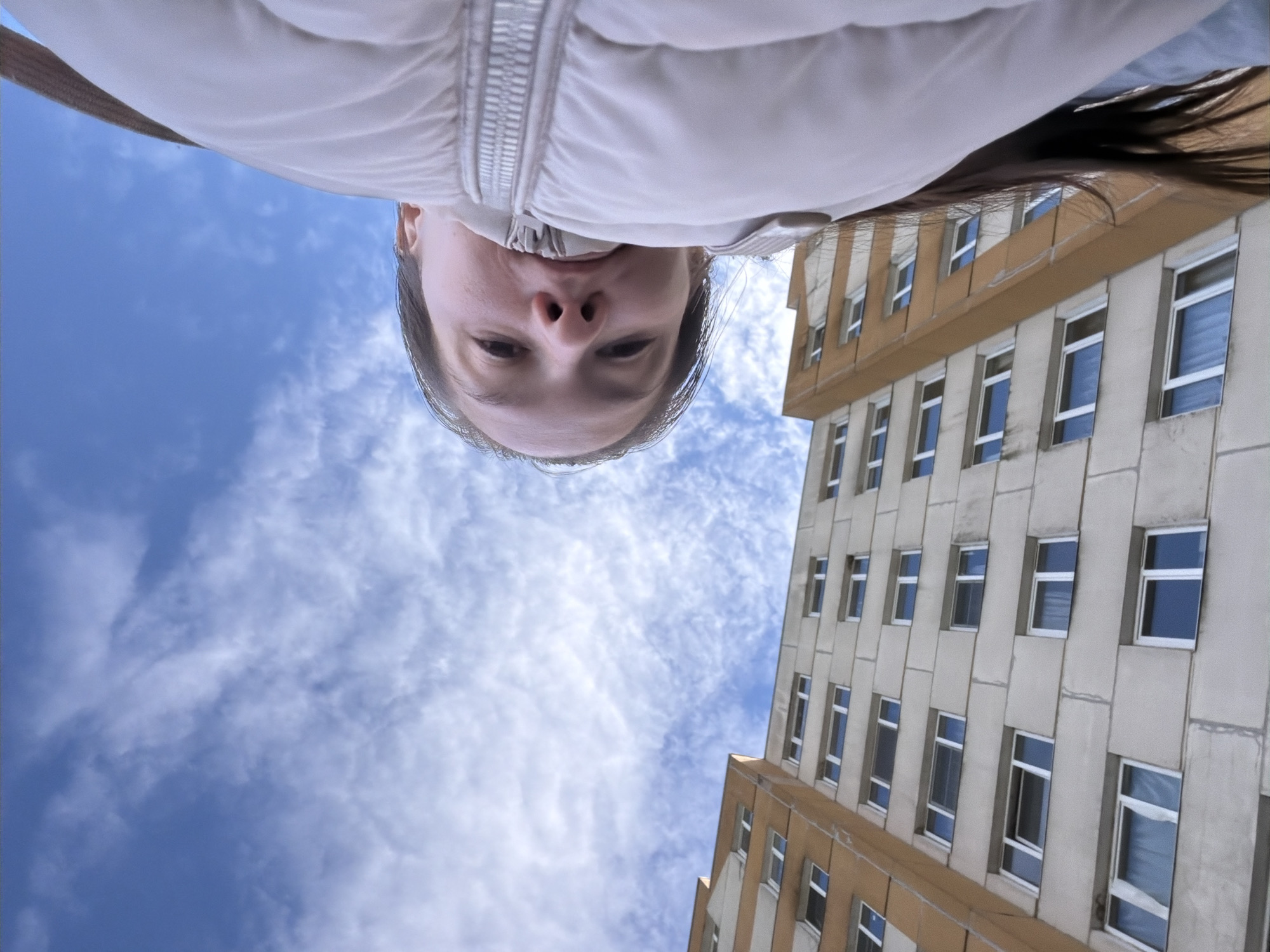 A person's face, cropped from the neck up, is positioned centrally in the frame. The person is wearing a light-colored puffer jacket. Behind the person, a building with multiple windows is visible. Beyond the building, the sky with scattered clouds serves as the backdrop. The location is potentially Staraya, Russia, based on the prompt.