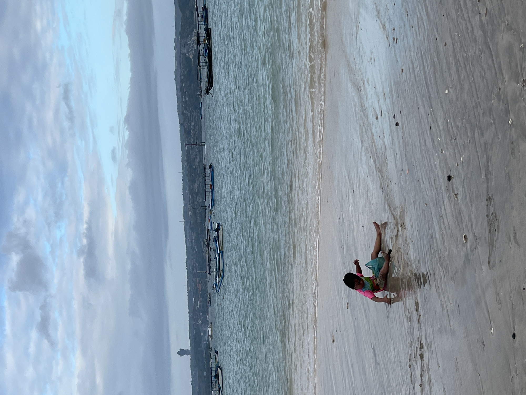A lone child, wearing a pink and green swimsuit, is sitting on the wet sand of Kuta Beach, Indonesia, reaching towards the water. The child has a turquoise bag next to them. The sea is calm with small waves lapping at the shore, and numerous boats are anchored in the distance. The sky is overcast with muted blues and grays. In the background, a hilly coastline with buildings and a prominent tower can be seen. The sand is damp and reflects the light.