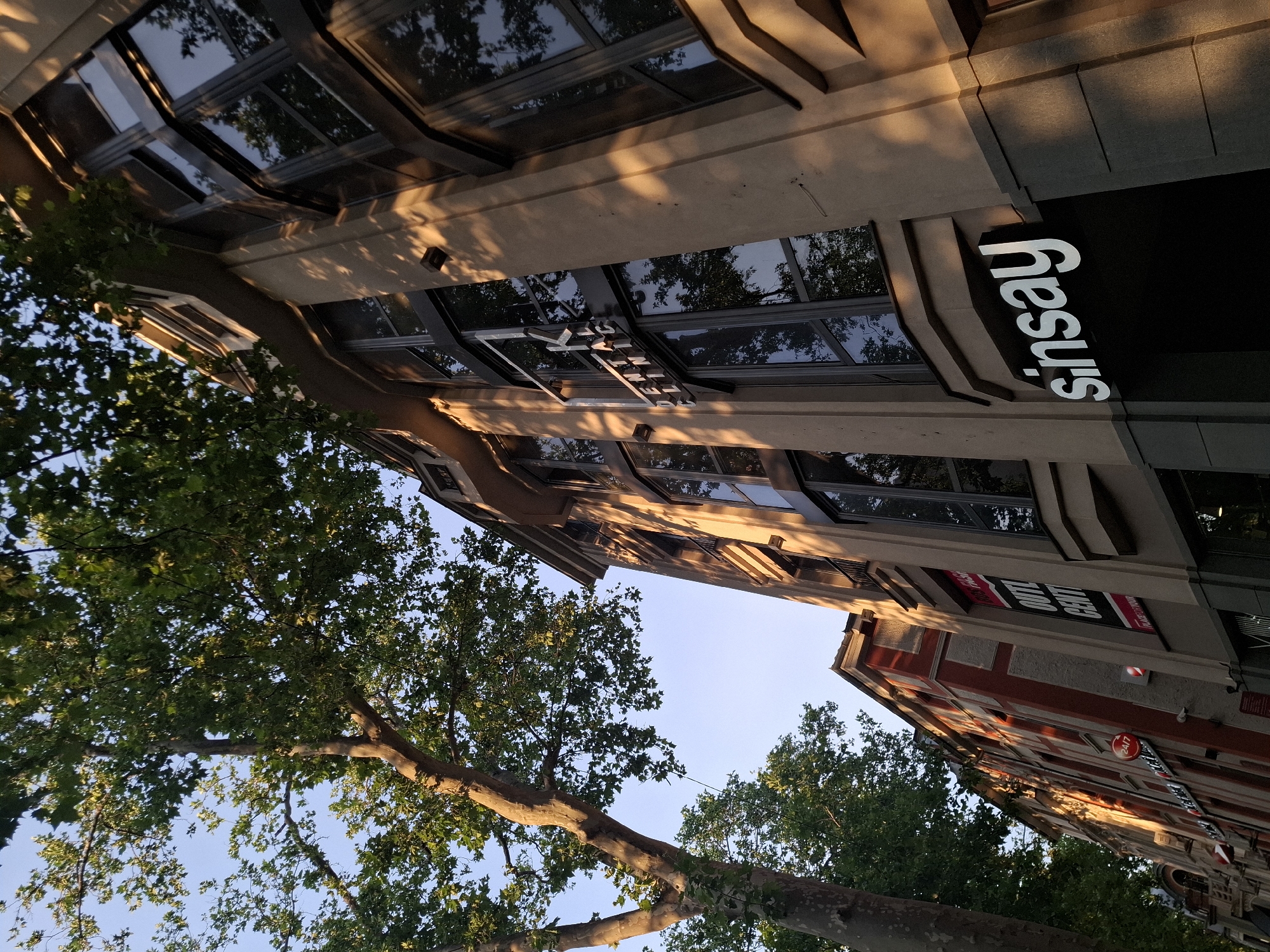 A low-angle shot captures a modern building with large windows and a beige facade on a sunny day in an urban setting. The building has multiple floors and is adorned with signage, including "K DENTAL CLINIC" and "Sinsay". To the left, a large tree with green leaves provides shade and is partially in sunlight. Another building with red and white accents is visible further down the street, also featuring signage like "OUTLET CENTER". The sky is clear and blue.