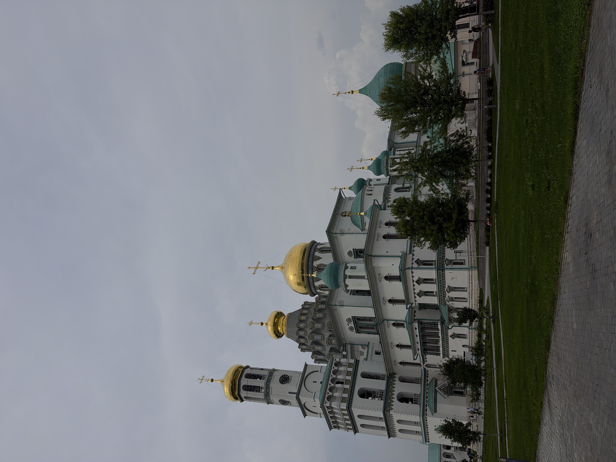 The image shows the Voskresensky Cathedral in Istra, Russia, under a cloudy sky. The cathedral is a large, white building with multiple gold and green domes, a tall bell tower with a clock, and intricate architectural details. Several trees are planted in front of the cathedral, and a paved walkway and grassy area are visible in the foreground. A few people are walking in the distance, and some are near the cathedral entrance. The overall scene depicts a serene and majestic religious complex.
