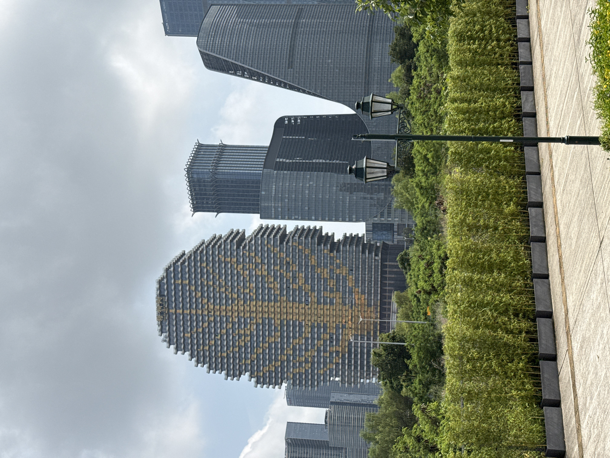 The image shows a view of several tall buildings in an urban area of Unknown, Macao. The buildings are modern in style, with the largest one being a high-rise shaped like an elongated oval with rows of alternating dark and light-colored sections. It appears to be a skyscraper with a repeating pattern of geometric shapes on its façade.
In the foreground, a row of green shrubbery runs parallel to a paved pathway. An old-fashioned street lamp is visible, with two lantern-style lights affixed to a dark green pole. The sky is overcast with light gray clouds.
The image presents a combination of natural elements (greenery, sky) with man-made structures (skyscrapers, street lamps, paved path). The buildings are of varying heights and designs, adding to the city's architectural diversity.