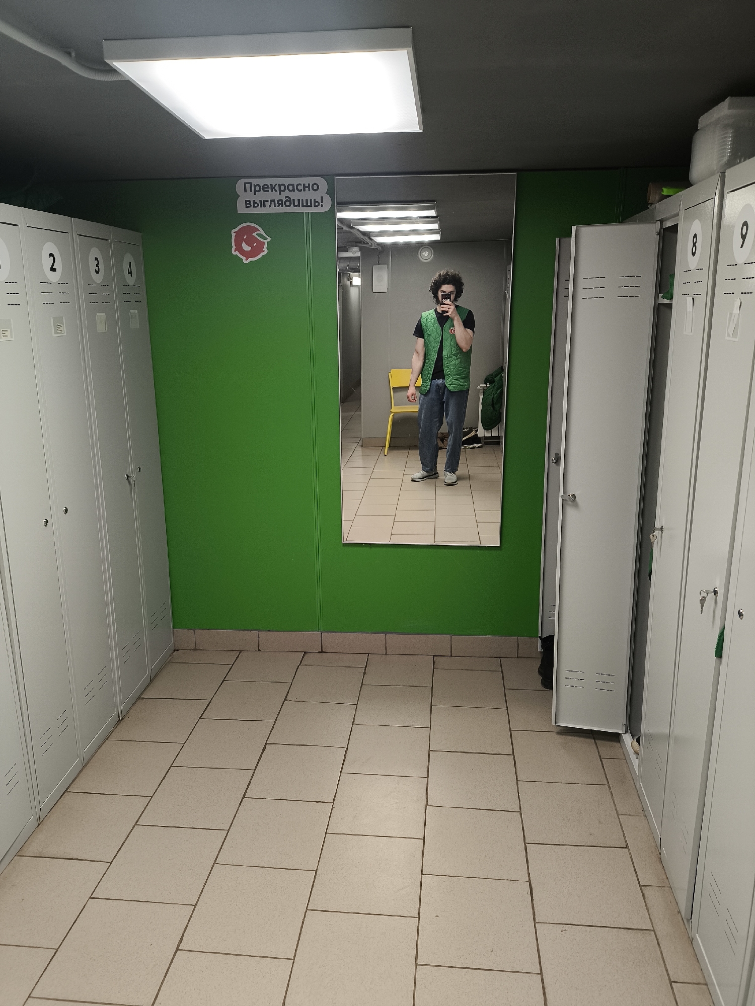 The image shows an interior space, likely a locker room. On the left and right sides of the frame are banks of metal lockers, numbered 1 through 4 on the left and 8 and 9 on the right. The walls are painted a bright green, and there is a rectangular mirror in the center of the frame. Reflected in the mirror is a person with curly hair, wearing a green vest, jeans, and a black t-shirt. The person is holding a phone and appears to be taking a photo. In the background of the mirror's reflection, there is a yellow chair and some other objects suggesting the locker room continues. The floor is tiled with light-colored rectangular tiles. On the green wall above the mirror, there is a sign in Russian that translates to "You look great!" along with a cartoon peach. The location is potentially in Chaikovsky, Russia.
