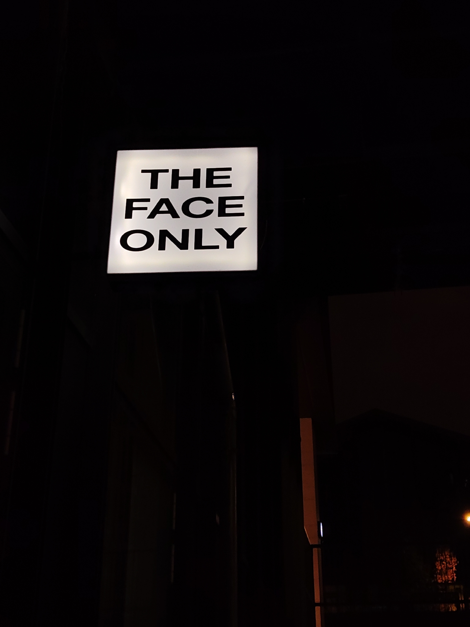 A low-angle, night shot focuses on a square, illuminated white sign attached to a dark structure, likely a building. The sign displays the black text "THE FACE ONLY" in a sans-serif typeface. The surrounding environment is mostly dark, with dim lighting revealing portions of the building, a window, and other architectural details. A vertical orange element is visible on the right side.
Given the unusual sign text, it's possible this image depicts a beauty salon or a similar establishment specializing in facial treatments. Without additional contextual clues, pinpointing a specific location in Moscow is not possible.