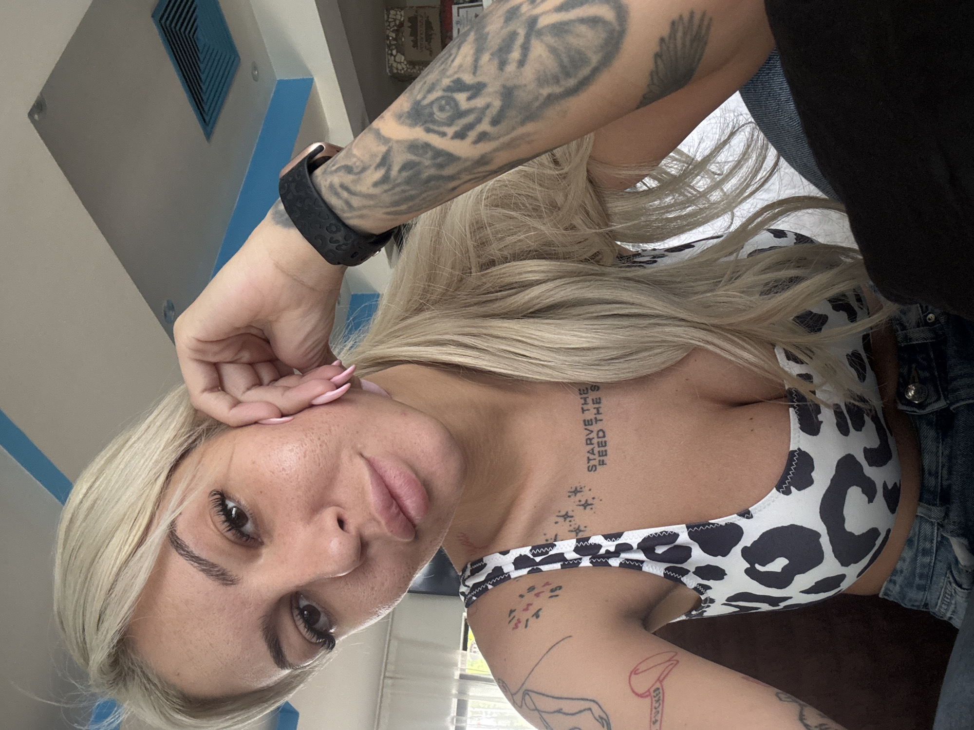 A blonde-haired woman with multiple tattoos is facing the camera. Her left arm, which is heavily tattooed, is raised to her forehead. She is wearing a smartwatch on her left wrist. She is wearing a cow-print top and a denim jacket. The background shows a wall with a blue and white air vent and other blurred items, possibly suggesting an indoor setting. The location in Plovdiv, Bulgaria cannot be determined from this image.
