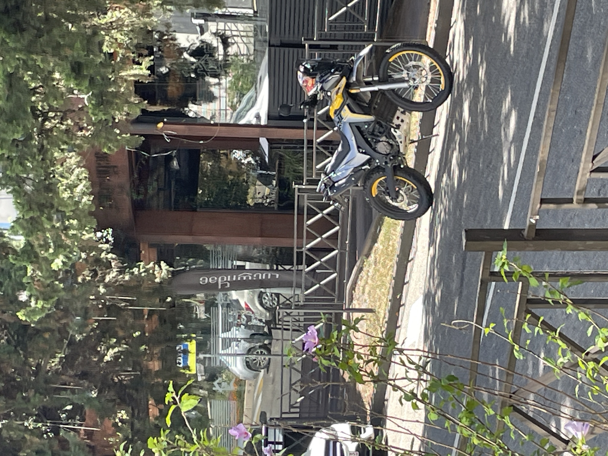 A silver and yellow dual-sport motorcycle is parked on a paved street in Sochi, Russia, under the dappled shade of trees. The motorcycle is positioned in the foreground to the right of the frame, facing left. It features a tall windscreen, knobby tires, and yellow accents on its wheels. Behind the motorcycle, a black metal fence with a geometric pattern runs horizontally across the middle of the image. Beyond the fence, a building with large windows and a dark wooden overhang is visible. In the background, a light-colored SUV is parked on a street, and a tall, dark sign with white text, which appears to be in Georgian script, is positioned near the building. Purple flowers are blooming on a branch in the lower left corner of the image, in front of the fence. The overall scene suggests a sunny day in an urban environment.