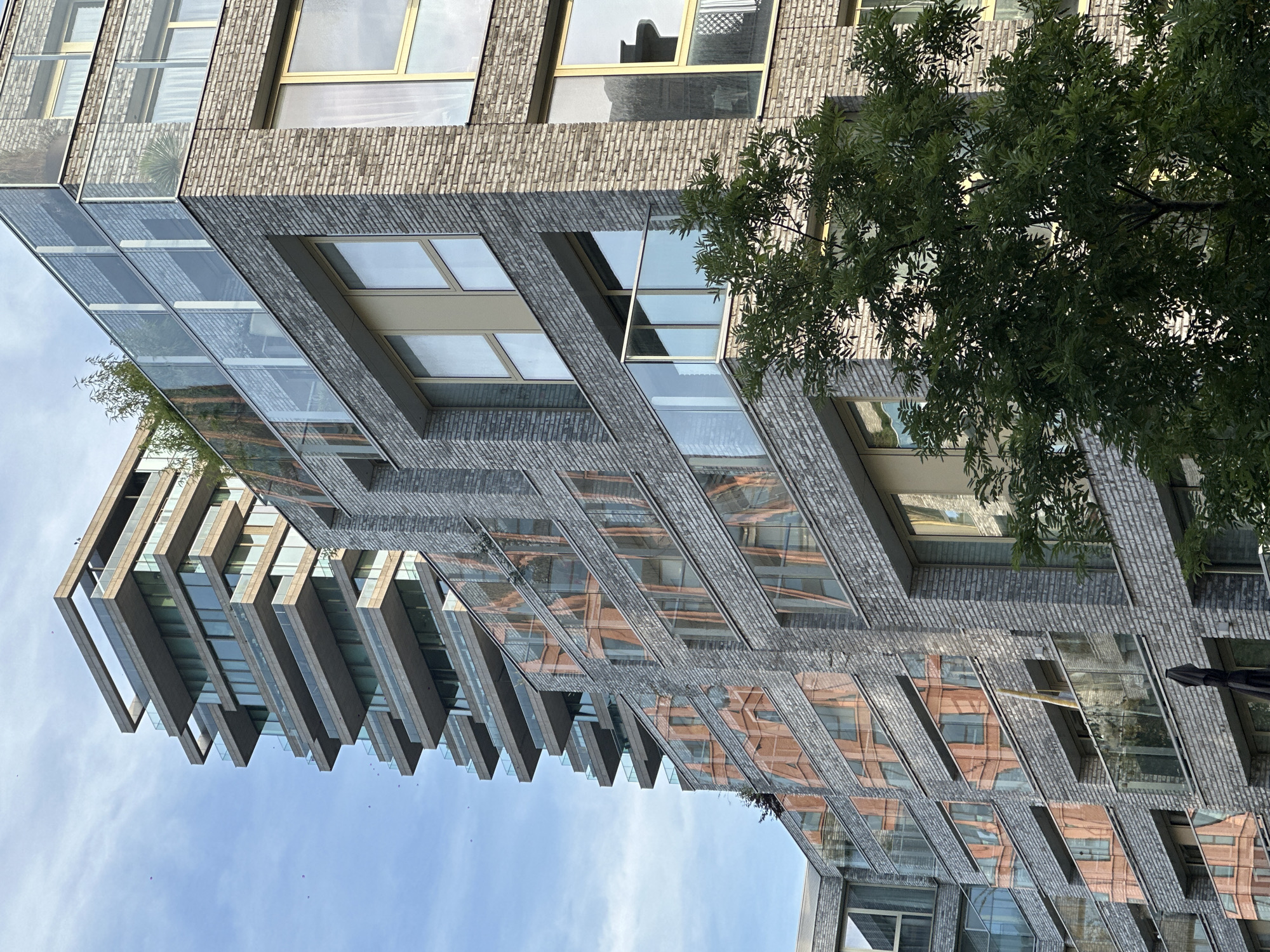 This image captures a modern architectural scene in Amsterdam, Netherlands, focusing on the interplay of contemporary buildings. In the foreground, a building with a grey brick facade features large windows with gold-colored frames and glass balconies. A lush green tree partially obscures the view of this building on the right side of the frame.
Behind and to the left, a taller building with a striking design is visible. This structure has multiple staggered balconies with glass railings, creating a layered effect. The facade of this building is composed of light-colored stone or concrete. The windows of both buildings reflect the surrounding environment, with the building on the left showing reflections of an orange-toned structure. The sky above is a pale blue with scattered clouds, and a few small birds can be seen in the distance. The overall impression is one of urban sophistication and modern design, set against a partly cloudy sky.