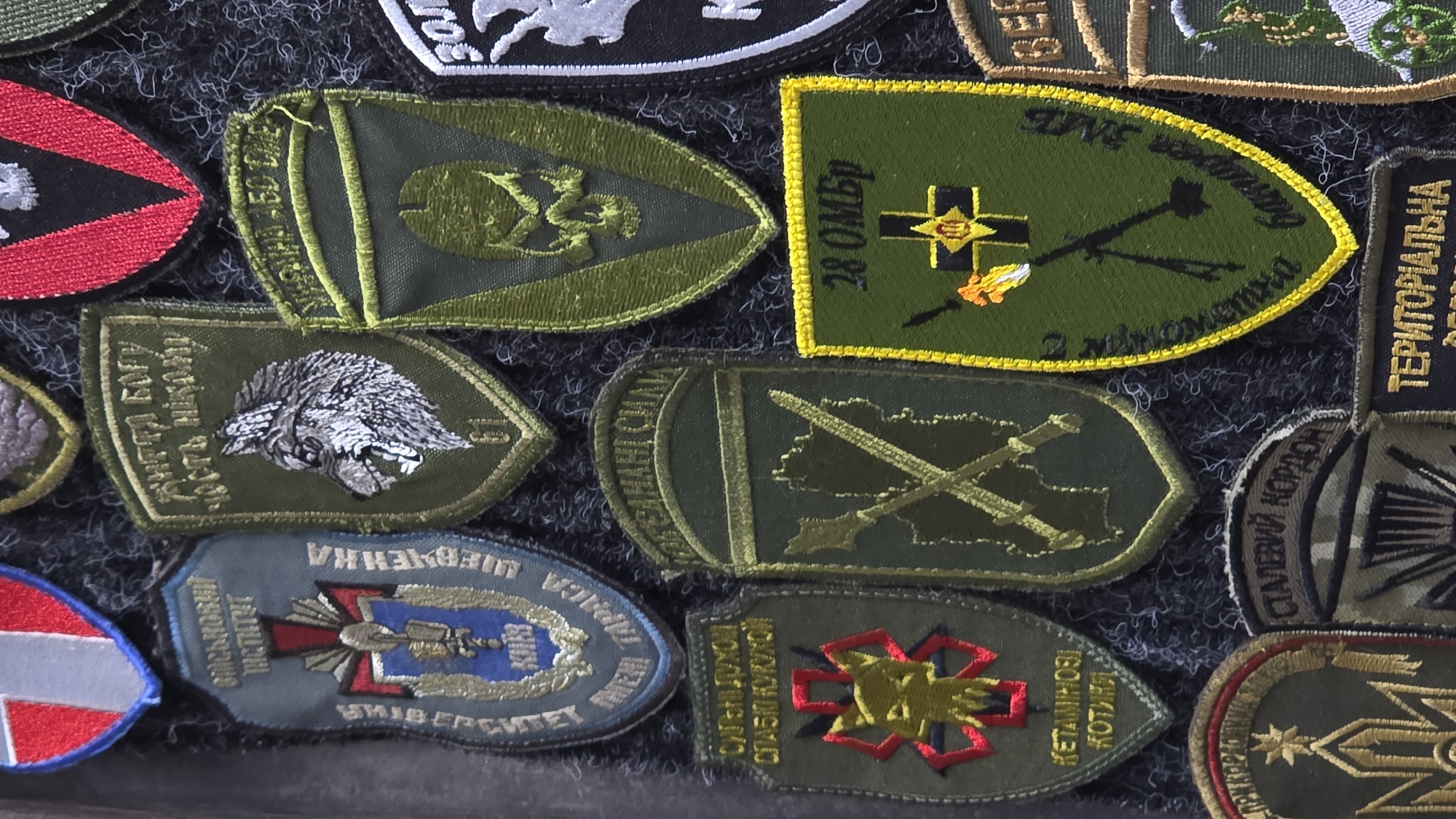 The image displays a collection of embroidered military patches, likely a display or a personal collection. The patches vary in shape and color, featuring military insignia, animals, skulls, and Ukrainian text. They are arranged on a dark, textured background, possibly fabric or felt. One prominent patch features a wolf's head with the text "ДИТЯ БОГУ, ЧЕСТЬ НІКОЛИ". Another shows a skull. A patch with the text "28 ОМБр" is visible. Other patches have designs of weapons and emblems. The overall impression is one of military memorabilia. There is no indication of the specific location within the city of Kyiv, Ukraine, nor any details about people, actions, or interactions, as the image focuses solely on the static display of patches.