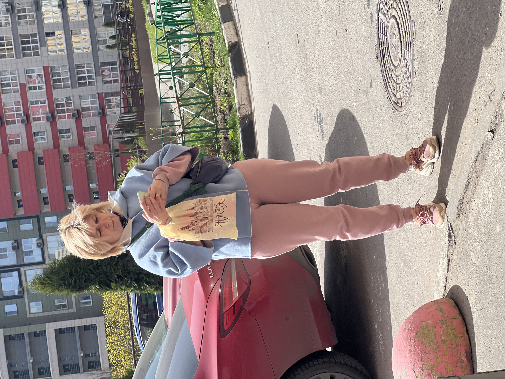 A woman with blonde hair stands outdoors in Khimki, Russia, next to a red car. She wears a light blue hooded sweatshirt, pink pants, and athletic shoes. She is looking at a small object she holds in her hands. In her other hand, she holds a tan-colored bag with the word "Хлеб" on it. Behind her is an apartment building with multiple windows and red accents. A green metal fence is visible. The ground is paved with asphalt. A round manhole cover and a red and pink painted concrete bollard are visible on the ground.