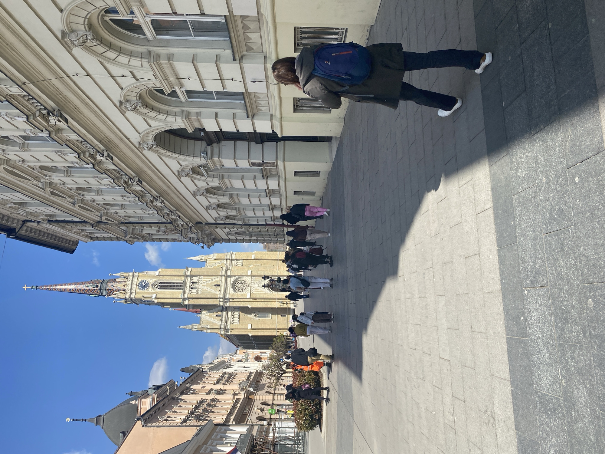 A medium shot shows an individual with a blue backpack walking along a paved street in Novi Sad, Serbia. The environment includes tall buildings with ornate architectural details, and a clear blue sky. Other people walk along the street, and the Matricaria Church is visible in the background.