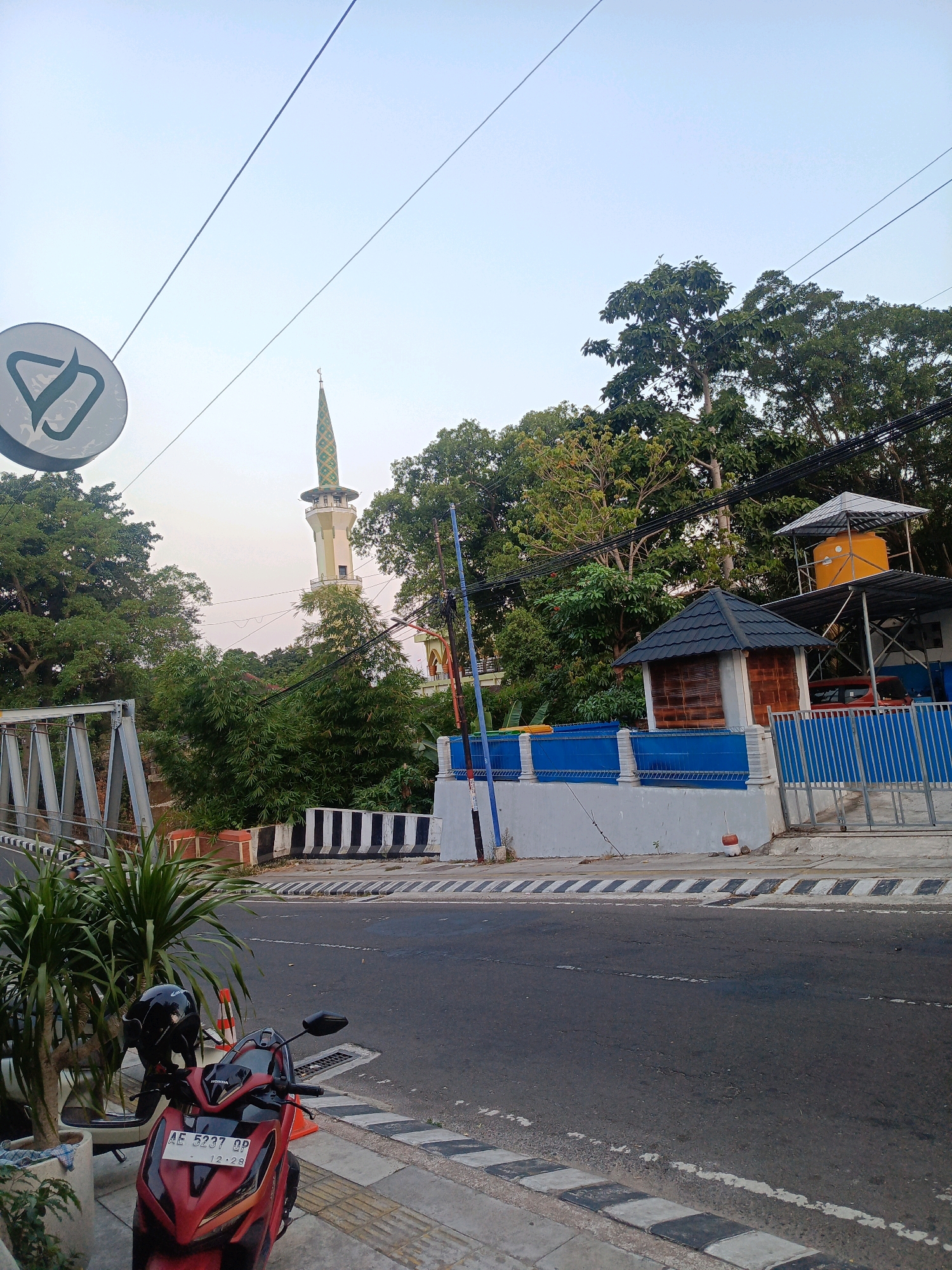 The image depicts a street scene in Magetan, Indonesia, likely in the afternoon or early evening. In the foreground, a red Honda motorcycle with license plate "AE 5237 QP" is parked on the sidewalk next to a potted plant. A black helmet rests on the motorcycle's seat. The street itself is paved, with clear lane markings and a pedestrian crossing marked with black and white stripes.
Beyond the street, a bridge structure is visible to the left. A prominent mosque with a tall, green and cream-colored minaret stands in the background, partially obscured by lush green trees. To the right of the mosque, there is a building complex with a blue fence and a small structure with a dark, tiled roof. Above this, a yellow water tank is supported by a metal frame. The sky is a pale blue, indicating daylight. The overall environment suggests a moderately developed urban area with a mix of infrastructure and natural vegetation.