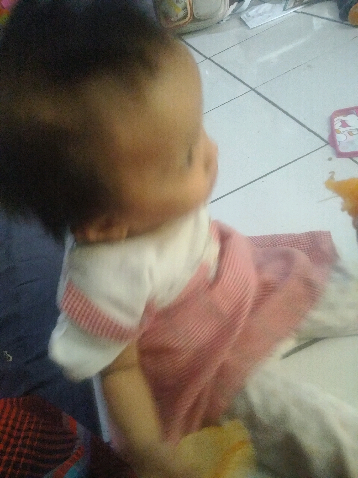 An infant is seated on a tiled floor in what appears to be a residential setting in Suka Sari, Indonesia. The infant is wearing a white and pink striped dress and is partially obscured by a blue fabric. The floor is white with visible grout lines. In the background, there are various objects, including what looks like a bag and a pink case with a cartoon character on it. The lighting suggests an indoor environment, possibly during the day or with artificial lighting. The focus is primarily on the infant, with the surroundings out of focus.