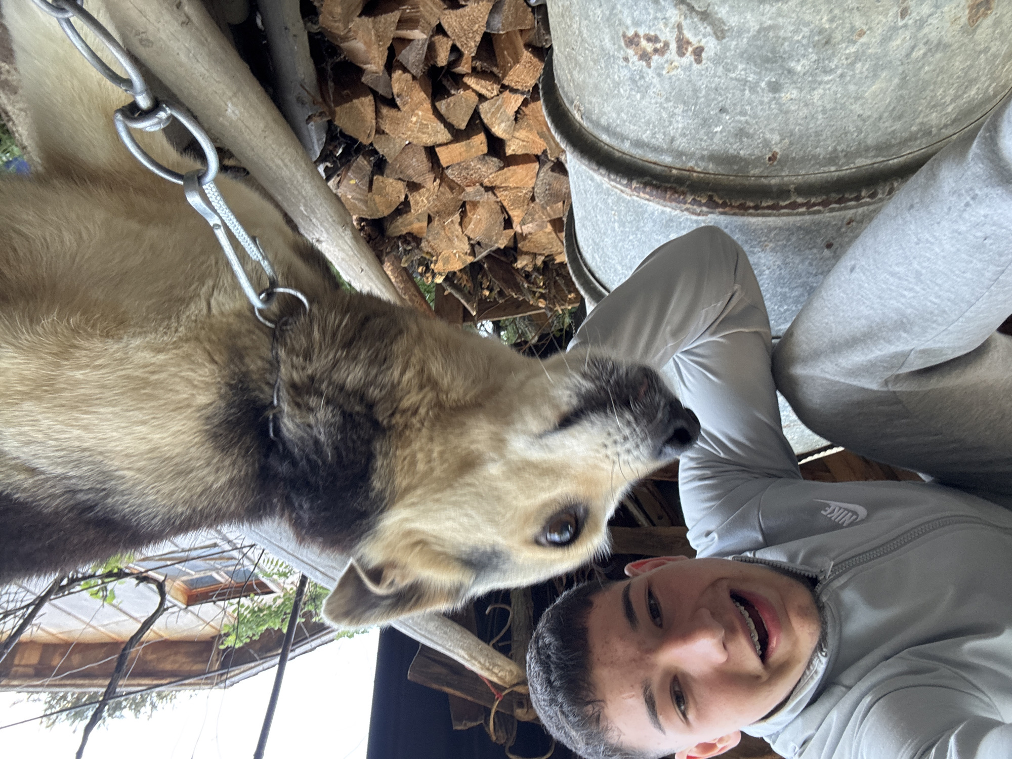 A young man is taking a selfie with a dog. The dog is leashed with a metal chain. Behind the dog and man are stacked firewood and a metal container. A wooden roof structure is partially visible in the background. The location is in a rural setting, presumably in or near Zlatograd, Bulgaria.