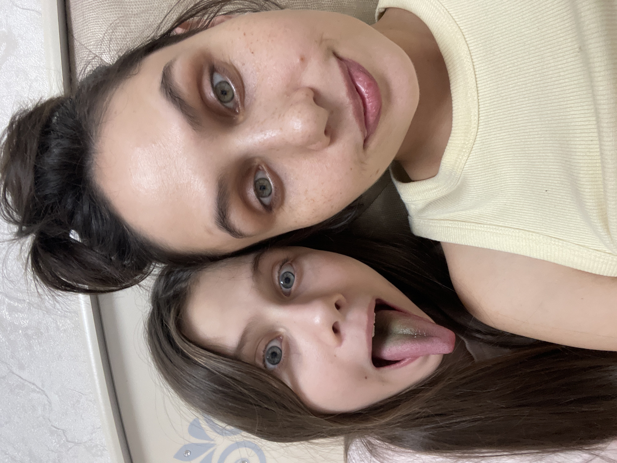Two females are in a close-up selfie-style shot, oriented on their sides. The female on top has dark hair pulled back in a bun, with a fair complexion, light green eyes, and is wearing a light yellow shirt. She is smiling slightly. The female on the bottom has long brown hair, light skin, and light blue eyes. Her mouth is open, and her tongue is sticking out, with a discolored greenish tint on the tongue's surface. The background appears to be a beige-colored wall or headboard. There is no location specified, and I am unable to discern whether the location is Izhevsk, Russia.