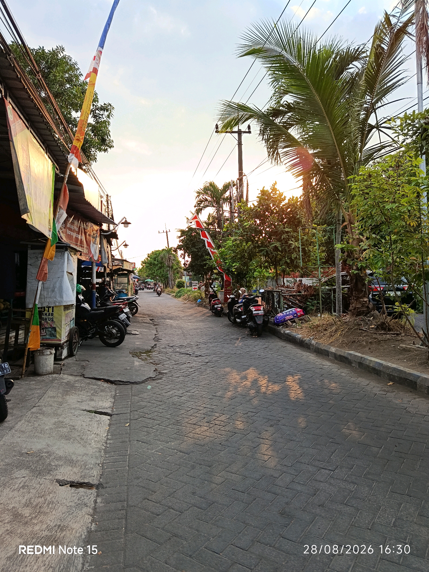 This image depicts a street scene in Keboansikep, Indonesia, likely during the late afternoon or early evening due to the lighting. The street is paved with interlocking gray bricks and has a slight incline. On the left side of the street, there are several small businesses or stalls with awnings and various signs. A prominent flag pole with a colorful flag, likely representing a local or national holiday, is visible. Parked along the left sidewalk are several motorcycles, a common mode of transportation in the area.
The right side of the street features a dense line of trees and vegetation, including a palm tree, with power lines and utility poles overhead. Sunlight filters through the trees, casting dappled shadows on the street. Further down the street, more trees and a few parked cars are visible. In the distance, a few people are seen on motorcycles, suggesting ongoing traffic. The overall atmosphere is one of a typical urban street in a tropical climate, with a mix of commercial activity and natural elements. The timestamp at the bottom indicates the image was taken on August 28, 2026, at 16:30.