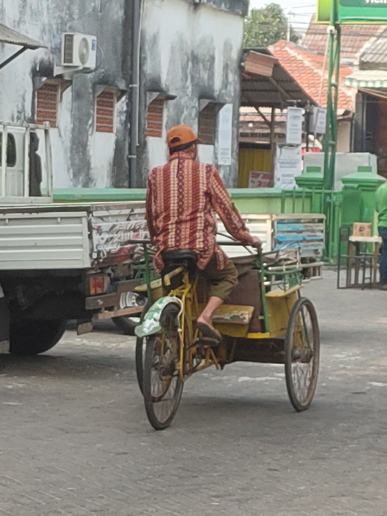 A person wearing an orange cap and a red and white patterned long-sleeved shirt is riding a yellow rickshaw. The rickshaw has three wheels, with the two larger wheels at the back and a smaller wheel at the front. The person is facing away from the camera, and their left leg is visible, wearing a sandal. The rickshaw appears to be carrying some items covered by a dark brown cloth.
In the background, a white truck with a silver side is parked. The truck has a green stripe along its side. Behind the truck and rickshaw, there are buildings with weathered, grey walls. Some windows have orange-red brickwork visible, and some have shutters or awnings. An air conditioning unit is mounted on one of the walls. Further in the background, more buildings with orange tiled roofs and green structures are visible. The scene appears to be on a paved street. The overall impression is that of a street scene in a bustling, possibly urban or semi-urban environment in Kediri City, Indonesia, suggested by the style of the buildings and vehicles.