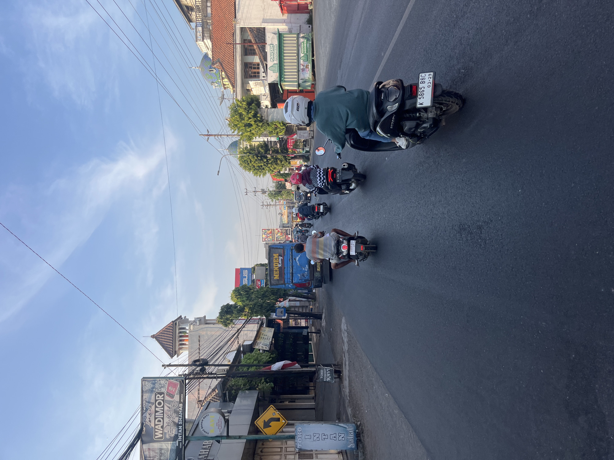 The image depicts a street scene in Kalinyamatan, Indonesia, likely during daylight hours. The perspective is from a moving vehicle, looking forward along a paved road. Several people on motorcycles are visible, navigating through traffic. On the left side of the road, buildings line the street, some with signs, including one for "WADIMOR." A blue truck with the word "MENDEM" on its side is also present in the traffic flow. The environment includes power lines overhead and some trees. The sky is blue with scattered white clouds. The overall impression is a typical urban or semi-urban street with active motor vehicle traffic.