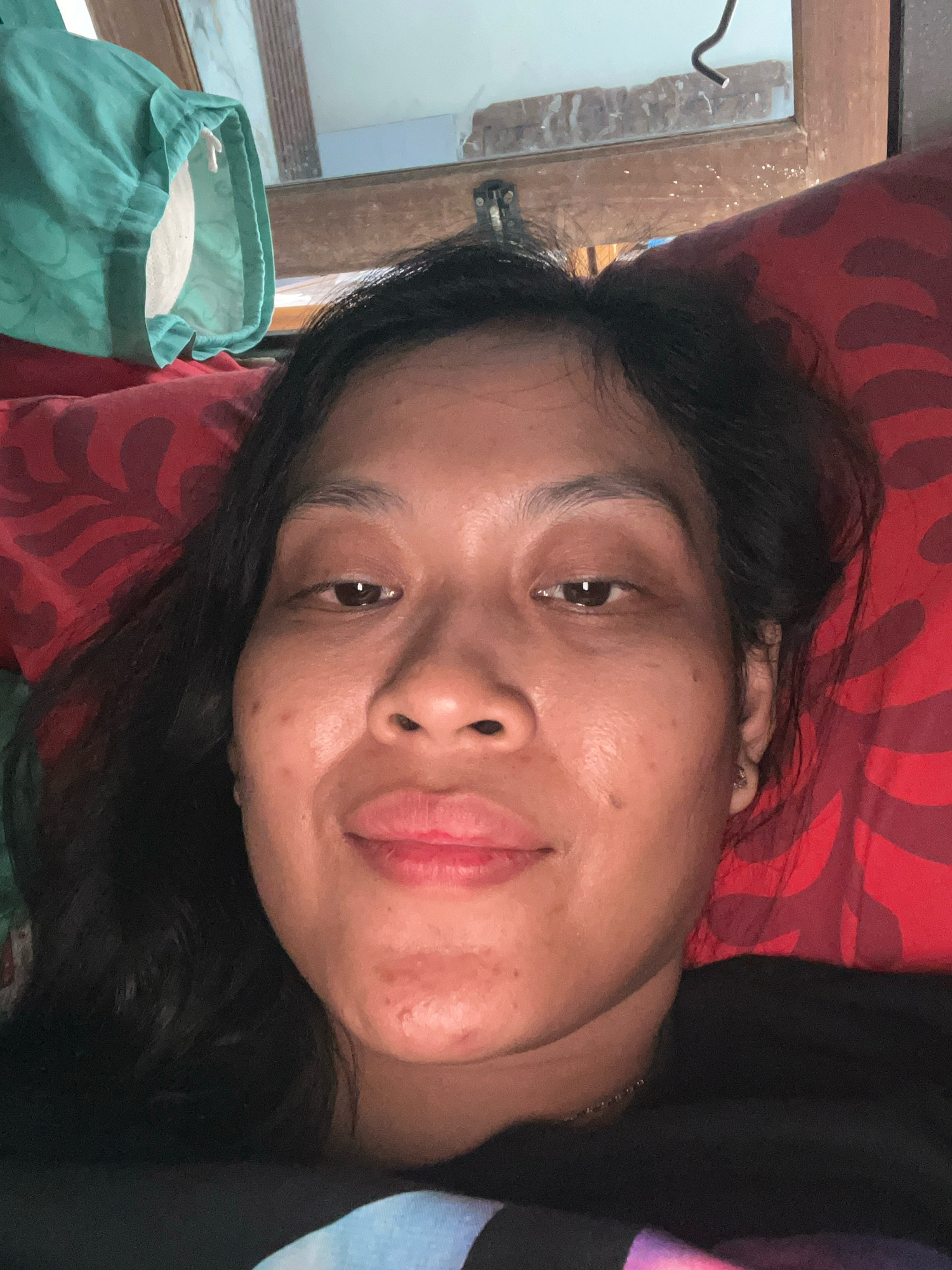 In Kecamatan Pati, Indonesia, a medium shot captures a woman lying down, her head resting on a red patterned pillow. She is facing the camera with her eyes open and a slight smile. Her dark hair is spread around her head. To the left of the frame, a teal fabric bag is visible, hanging from a wooden frame. The background shows a mirror reflecting part of the ceiling and a wooden structure. The woman is wearing a black t-shirt.