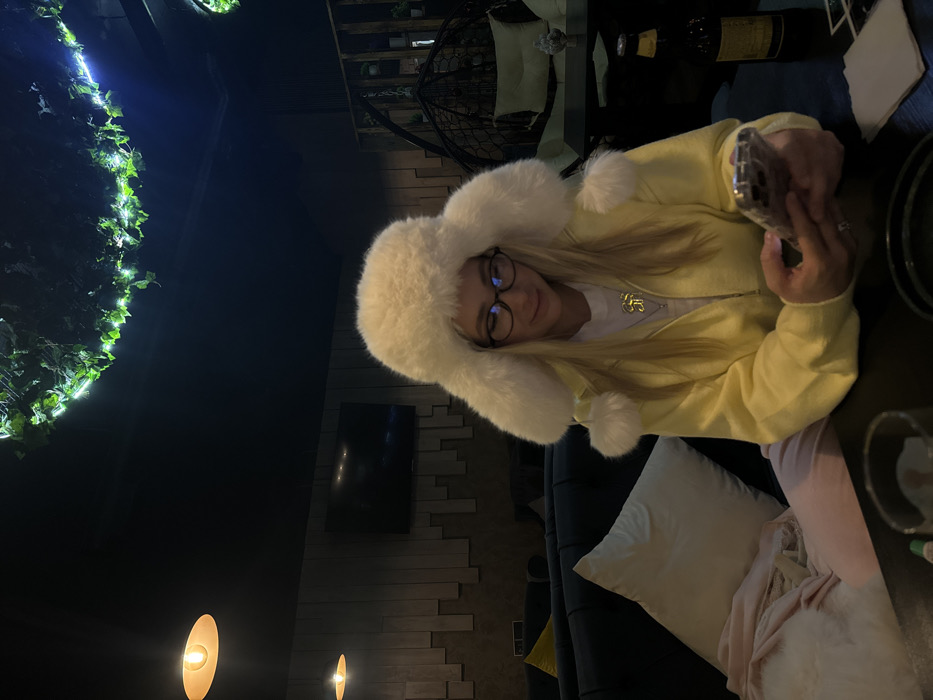 A woman with blonde hair wearing glasses and a large white fur hat is seen taking a selfie. She is wearing a yellow cardigan over a white shirt with a gold pendant necklace. She is sitting at a table with a wine bottle and glasses. A white pillow and pink blanket are visible on the couch she is sitting on. In the background, there is a decorative green light fixture and a wall with a television.