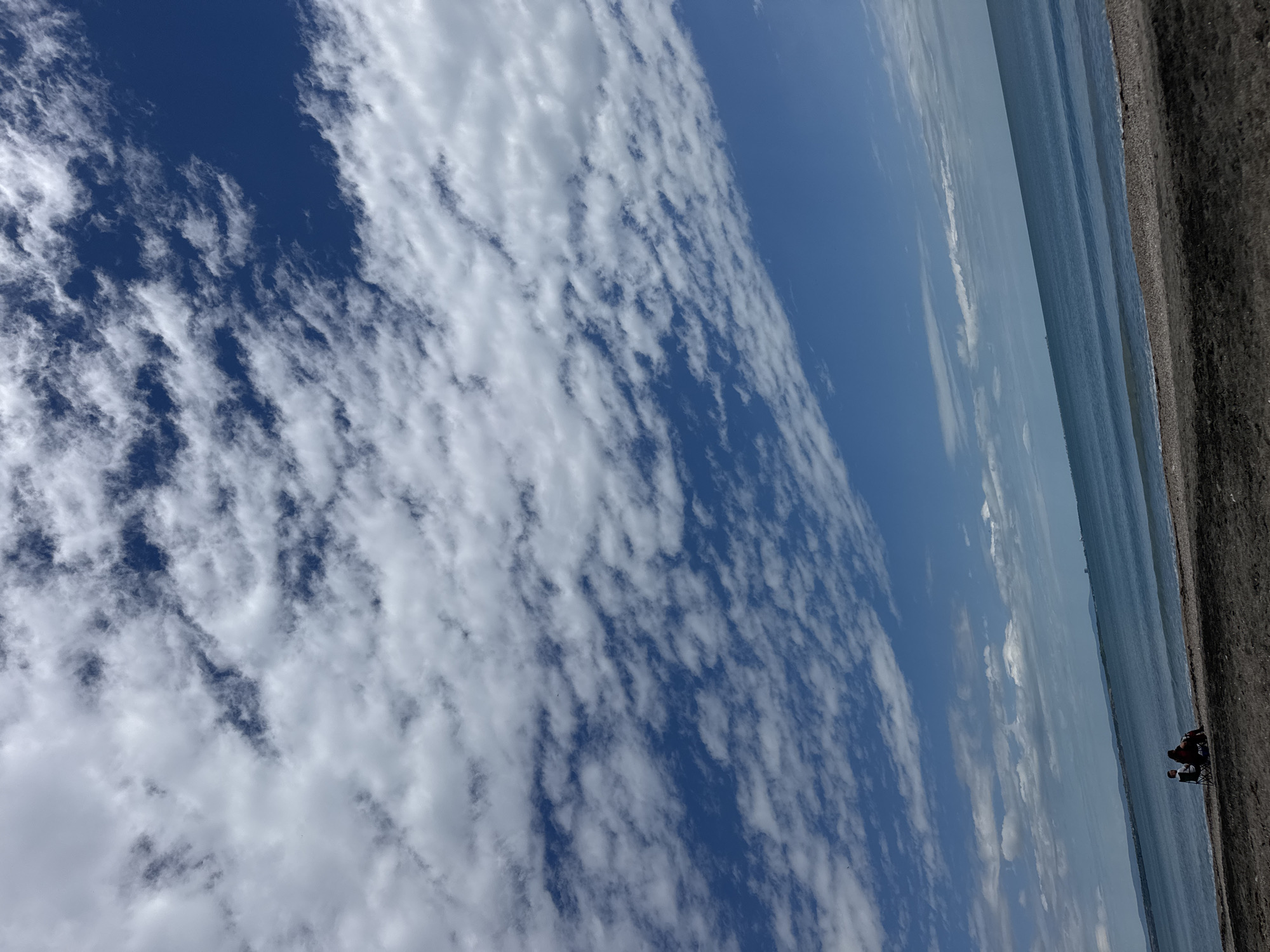 The image shows a coastal scene in Burgas, Bulgaria. The view encompasses a bright blue sky filled with elongated altocumulus clouds, transitioning to the sea and a dark, gravelly beach. The shoreline is nearly vertical in the image's perspective. Towards the bottom right, two individuals are visible, appearing to be seated near each other on the beach.