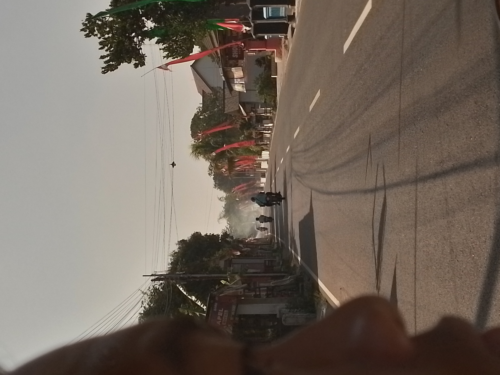 The image depicts a street scene in the city of Ngancar, Indonesia. The perspective is from a vehicle, with a portion of a person's face or body out of focus in the foreground left. The street is paved with asphalt and has white lane markings. Several motorcycles are visible on the road, with one in the foreground on the right appearing to be the subject of the shot, its rider leaning into a turn. A trail of smoke or dust is behind another motorcycle further up the road.
Along the sides of the street, there are buildings and trees. Red and green flags are strung along poles on the right side of the road, and some are also visible on the left. The buildings on the left include a shop with signage that reads "BARU APEL ROYAL" and some text below it. On the right side, there is a building with signage that reads "SHOP". Power lines are strung overhead across the scene. The sky is clear and overcast, suggesting it is daytime. The overall impression is a bustling street scene in a populated area of Ngancar.