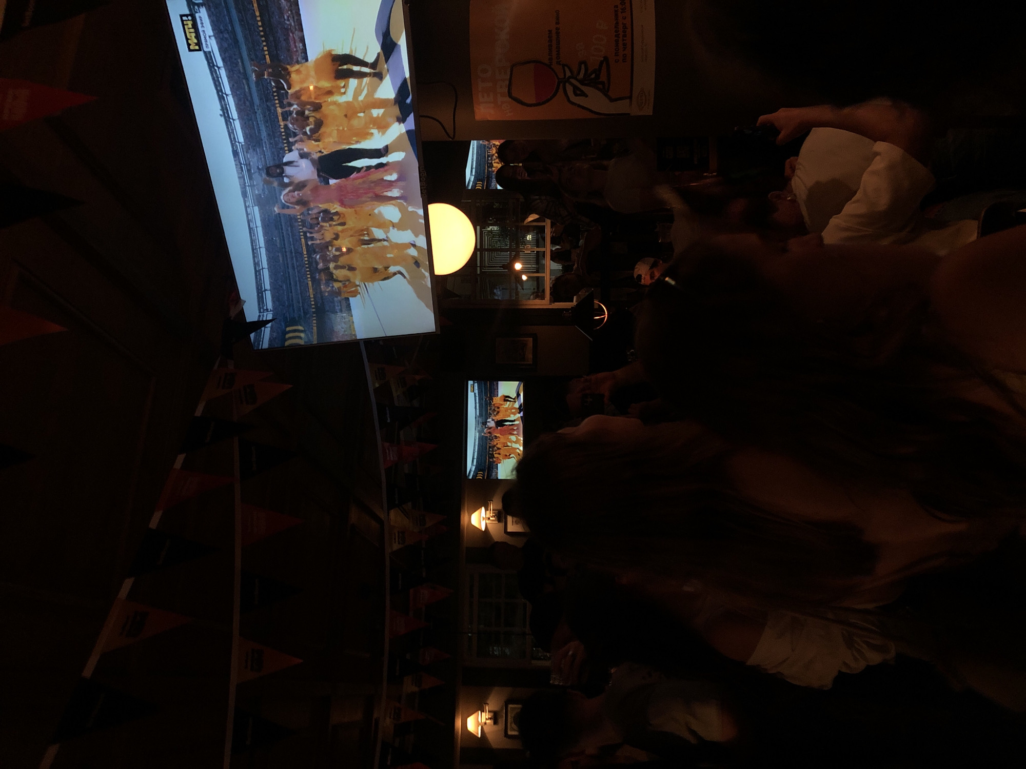 The image depicts a dimly lit interior of what appears to be a bar or restaurant, with a crowd of people in the foreground, largely silhouetted against the ambient light. In the background, a large screen displays a performance, possibly a concert or sporting event, featuring several individuals dressed in vibrant yellow attire and a woman in a pink dress. The screen also shows a logo that says "МАТЧ!" which is a Russian sports television channel. There are decorative triangular flags hanging from the ceiling, predominantly in orange and dark colors, suggesting a festive or event-themed atmosphere. Two wall-mounted lamps with conical shades provide a warm glow on the left side of the image. To the right, a poster with a stylized image of a hand holding a wine glass and Russian text is visible. The overall impression is of a lively gathering watching a broadcast event in a public venue in Moscow, Russia.